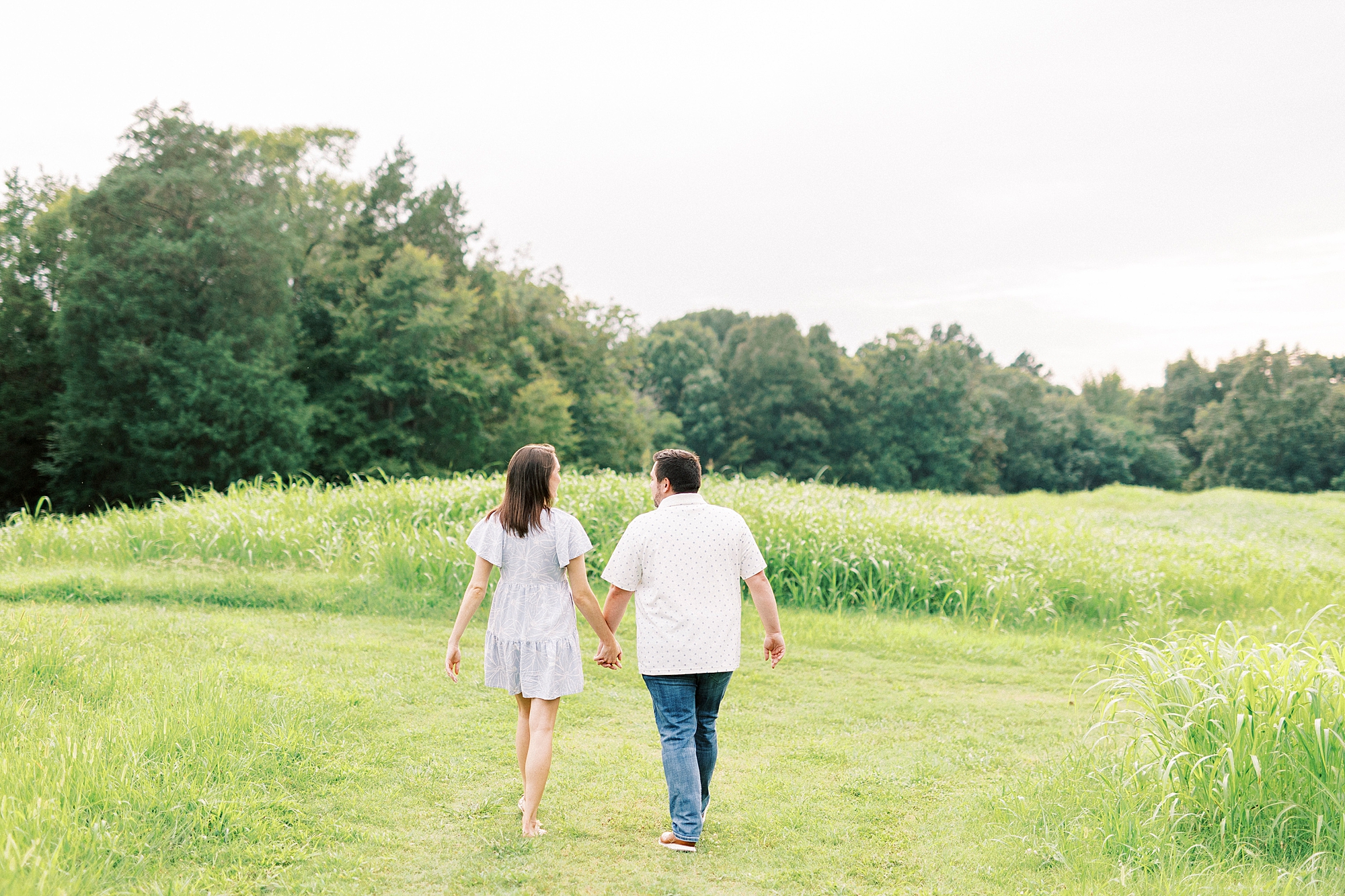 man and woman hold hands walking towards field during Kannapolis NC engagement session
