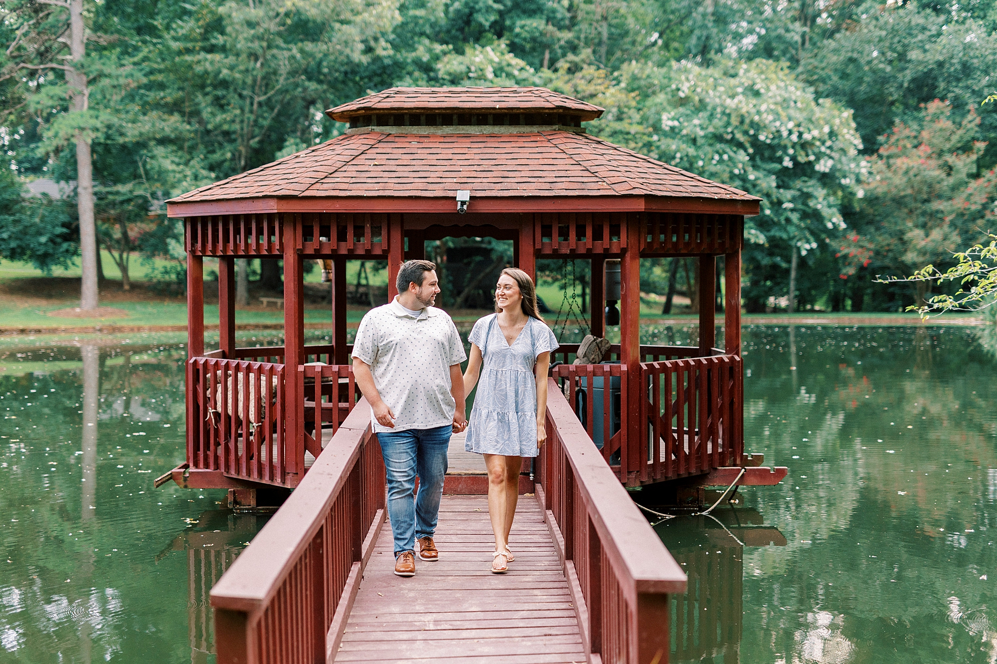 engaged couple smiles walking down wooden path near gazebo