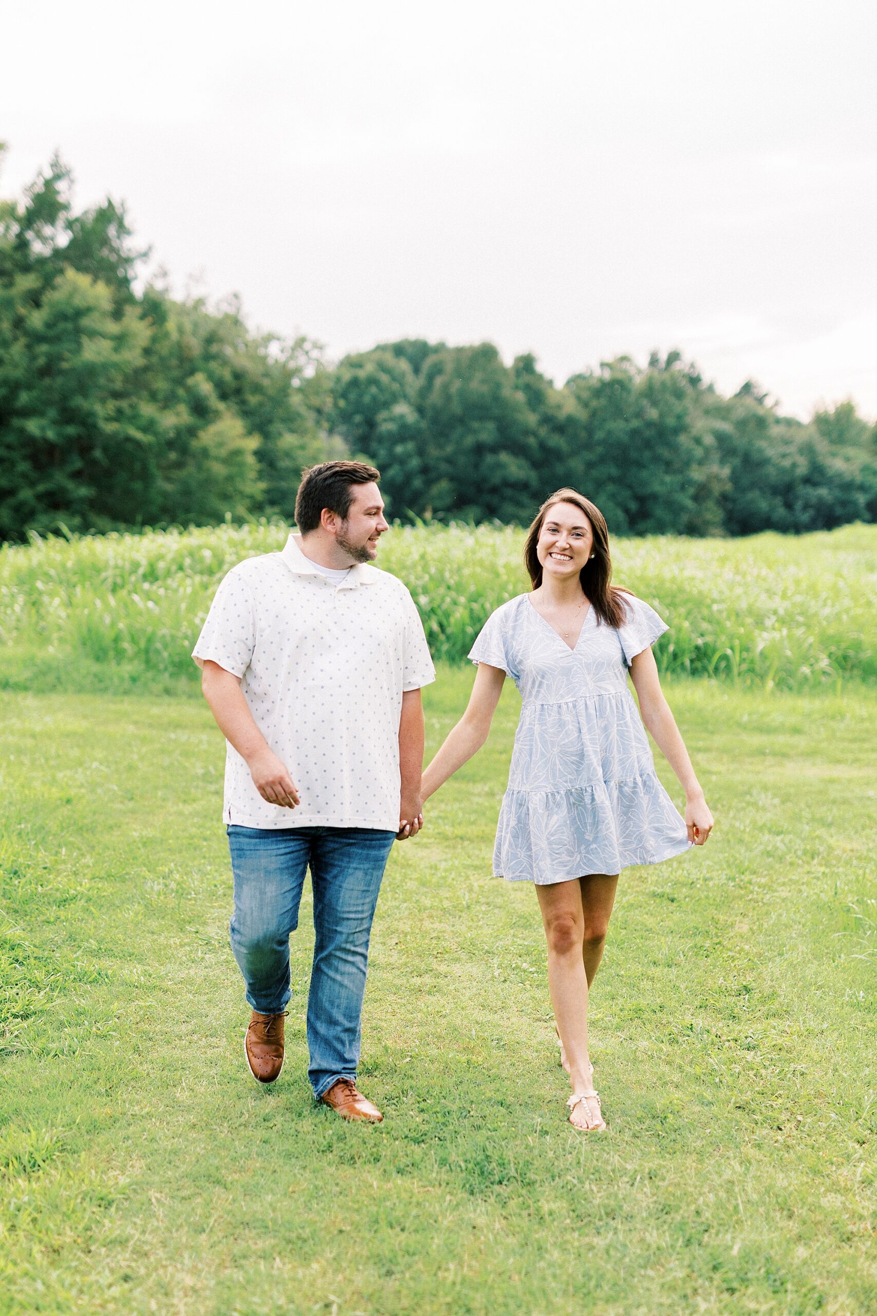 engaged couple holds hands walking away from field