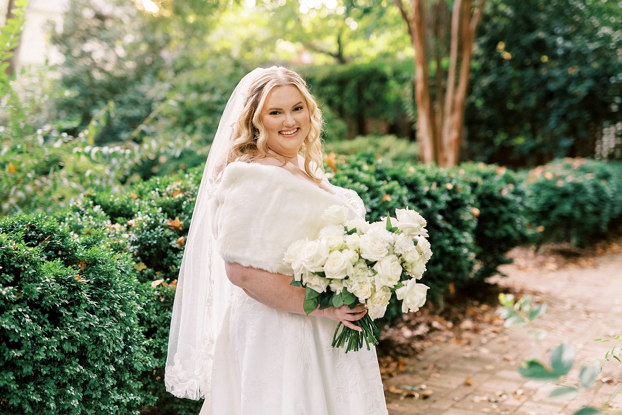 bride smiles with fur wrap holding bouquet of ivory roses inside Wing Haven Gardens