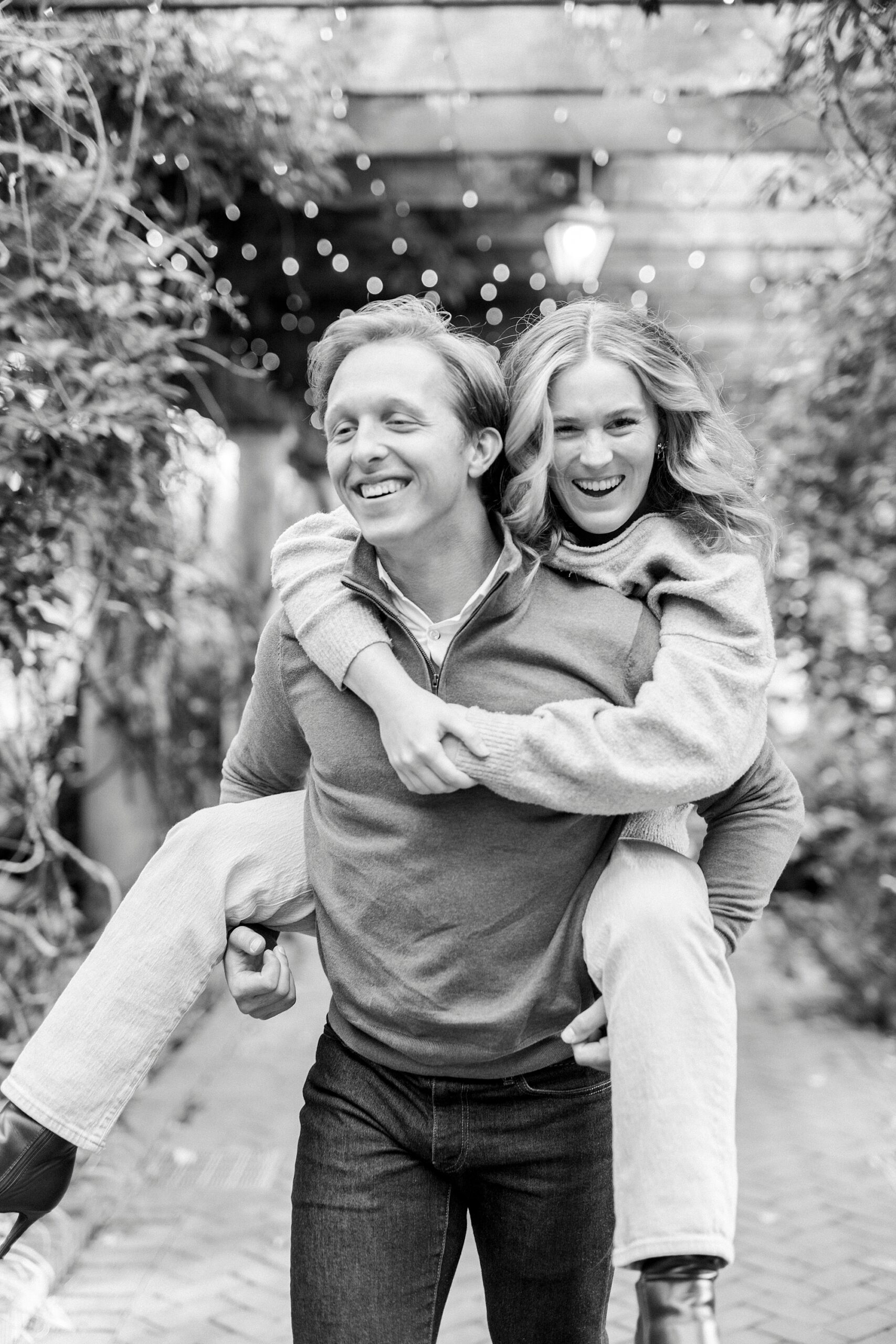 man gives woman piggy back ride during engagement photos inside Daniel Stowe Botanical Garden