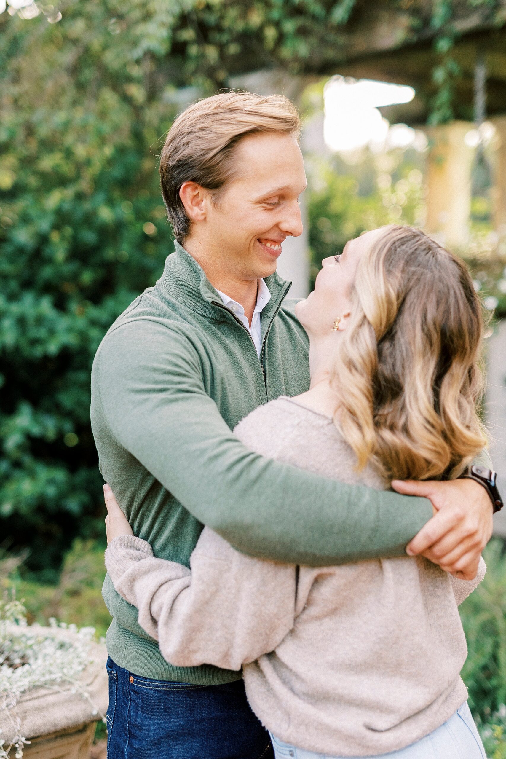 man in green sweater hugs fiancee during engagement photos at Daniel Stowe Botanical Garden