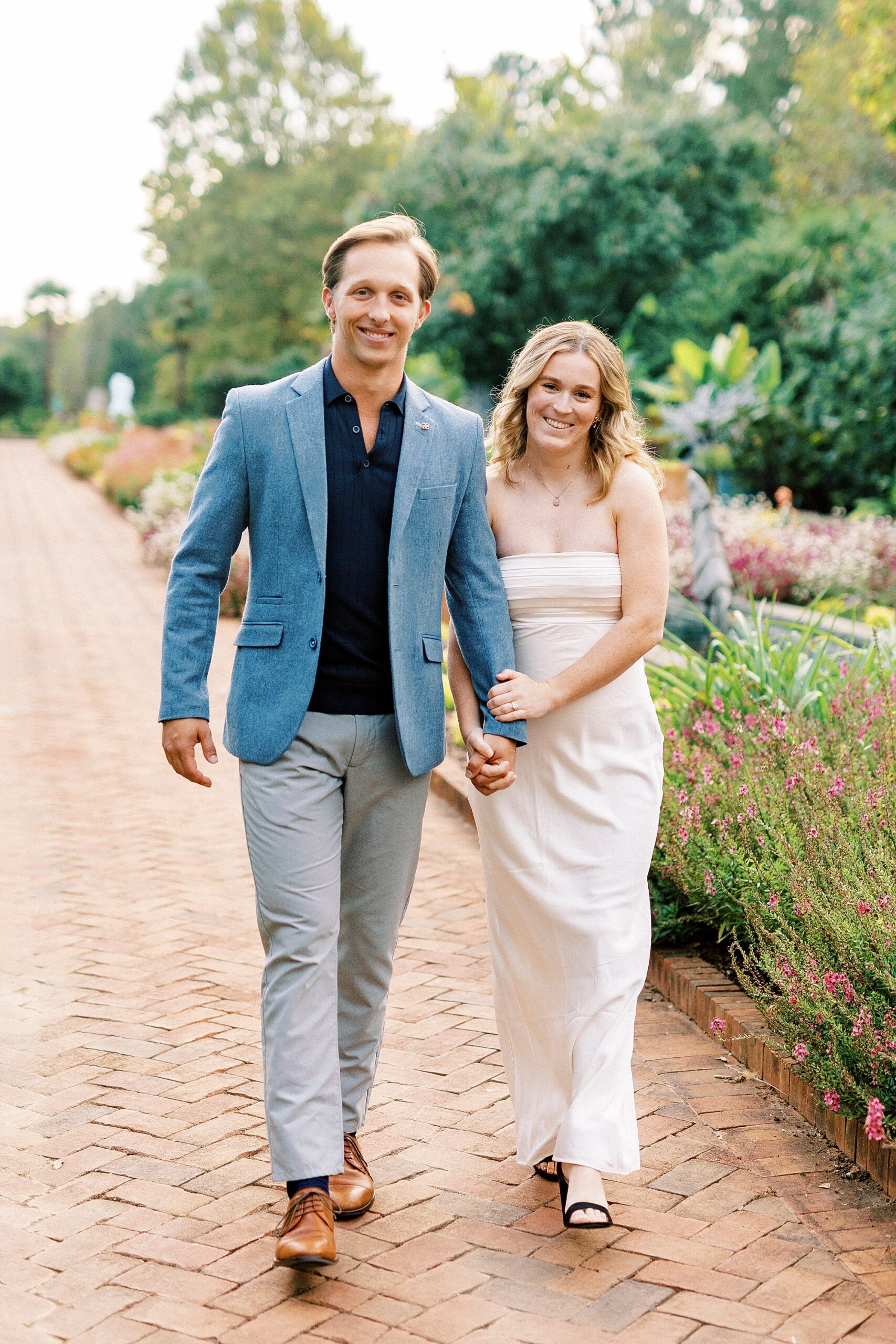 engaged couple holds hands walking on brick pathway in gardens