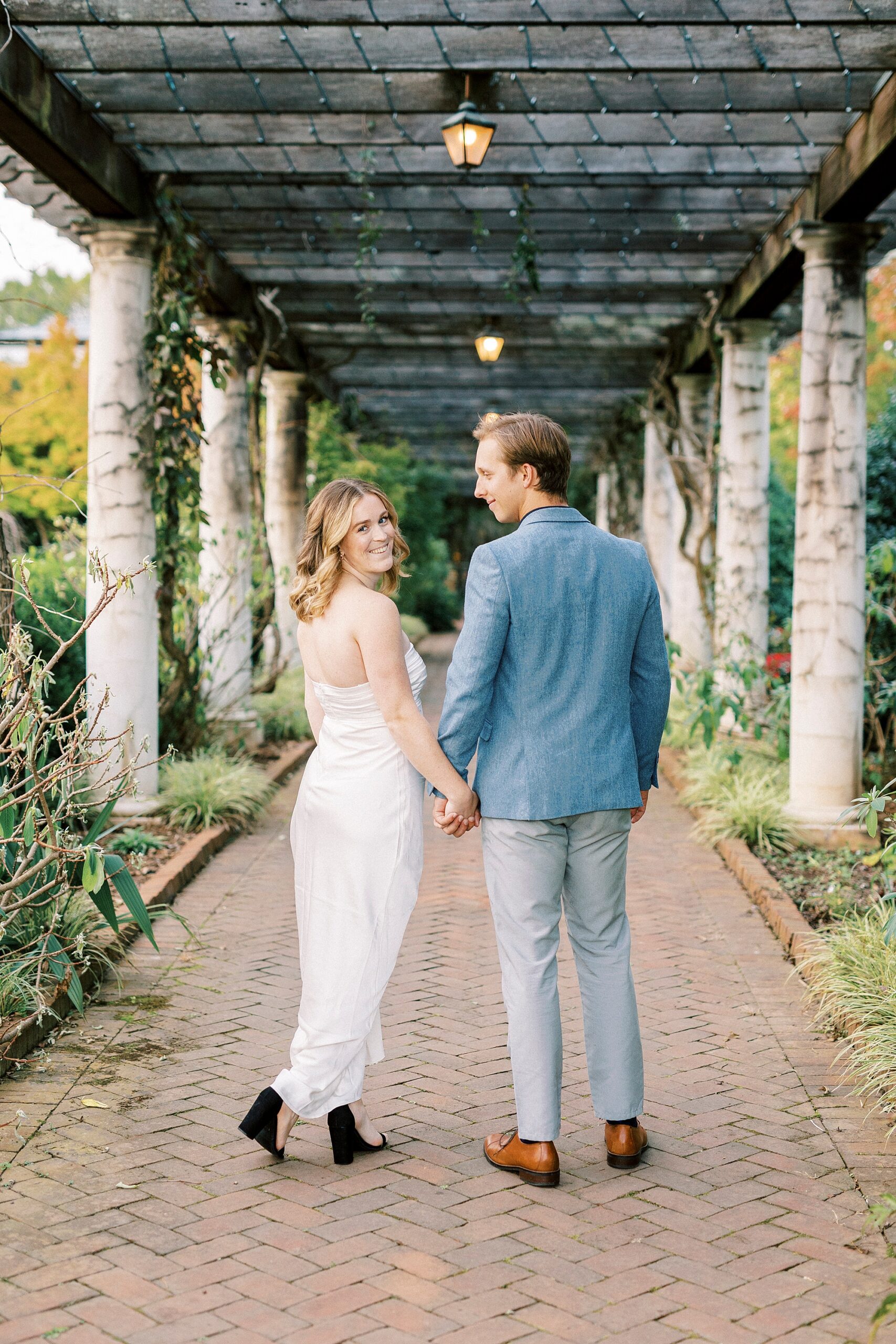 couple holds hands walking down pathway while bride looks over her shoulder 