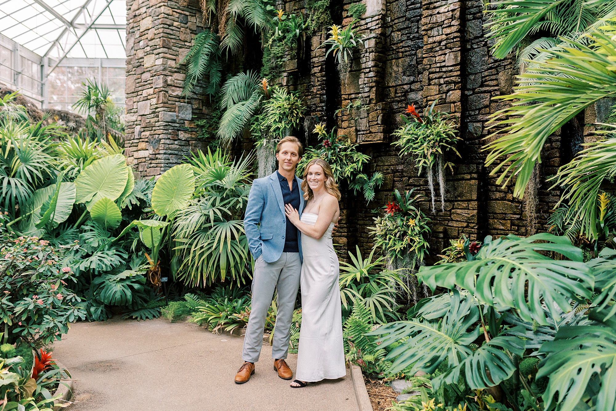 engaged couple hugs near stone wall inside Daniel Stowe Botanical Garden tropical display
