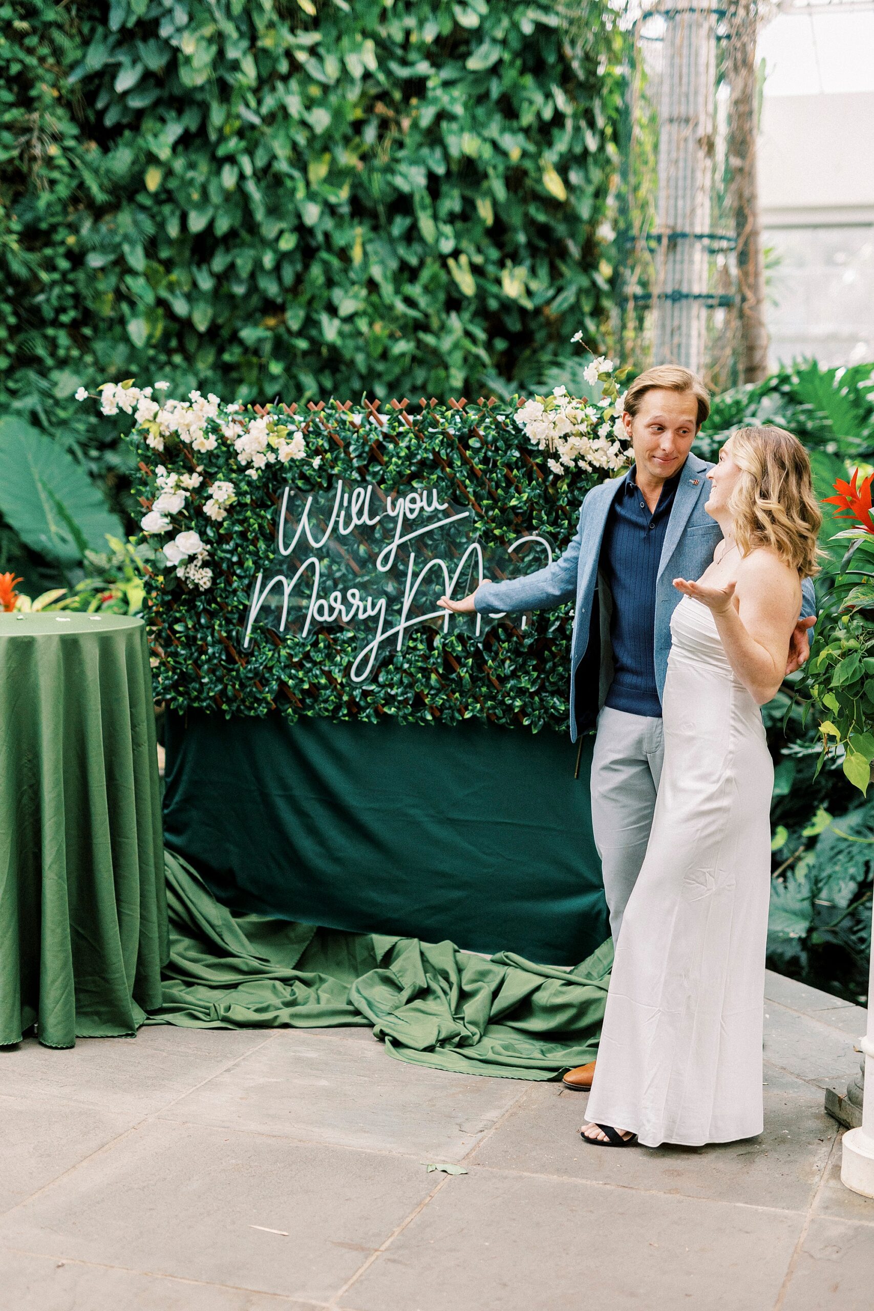 engaged couple laughs shrugging pointing to neon sign that says "will you marry me?"