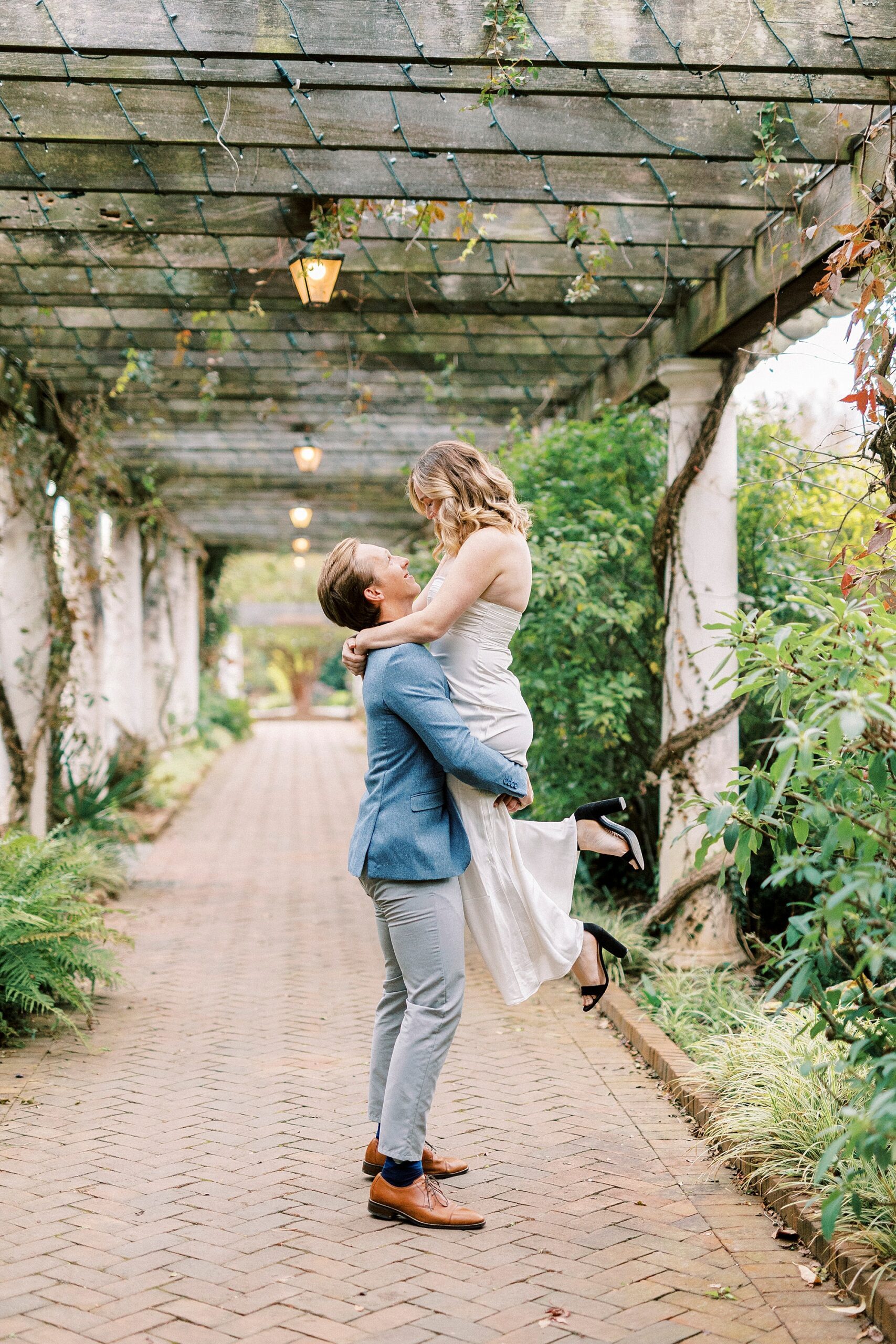 man lifts up woman in white dress during engagement photos in garden
