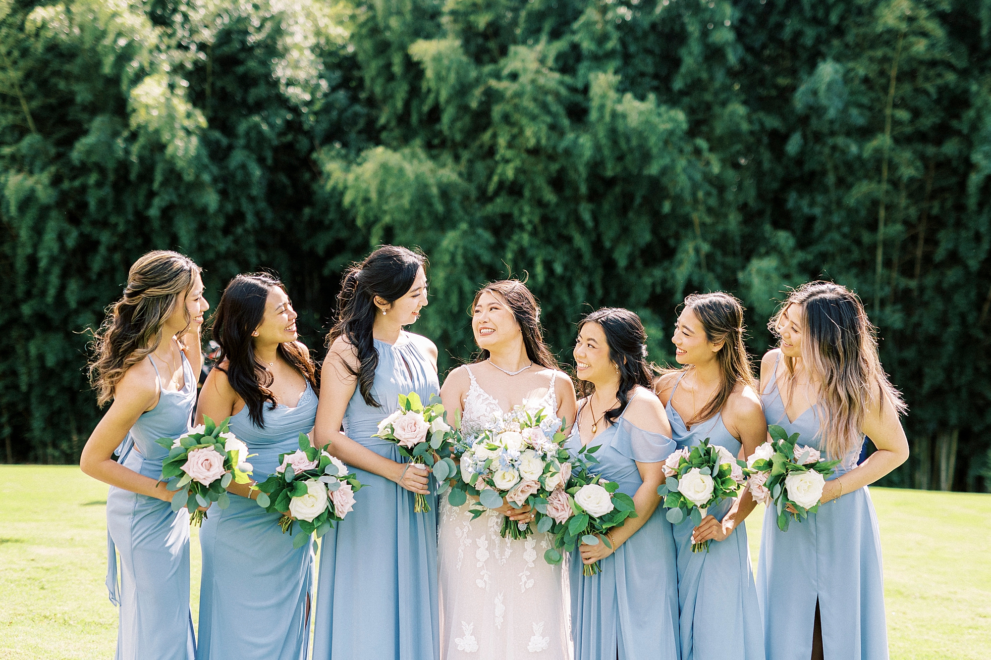 bride smiles at bridesmaids in light blue gowns on lawn at Camelot Meadows