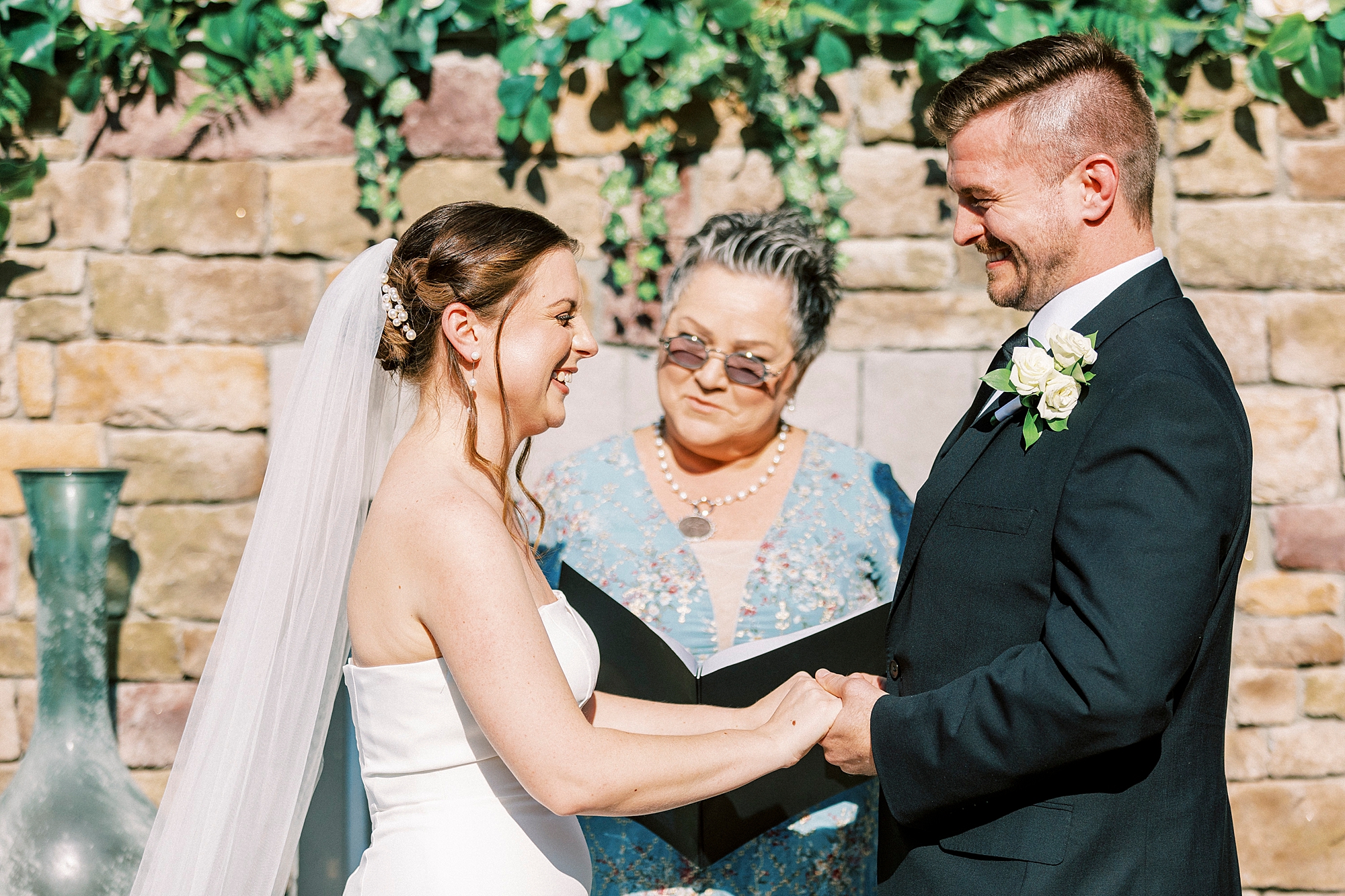 bride and groom smile holding hands during ceremony in front of brick fireplace