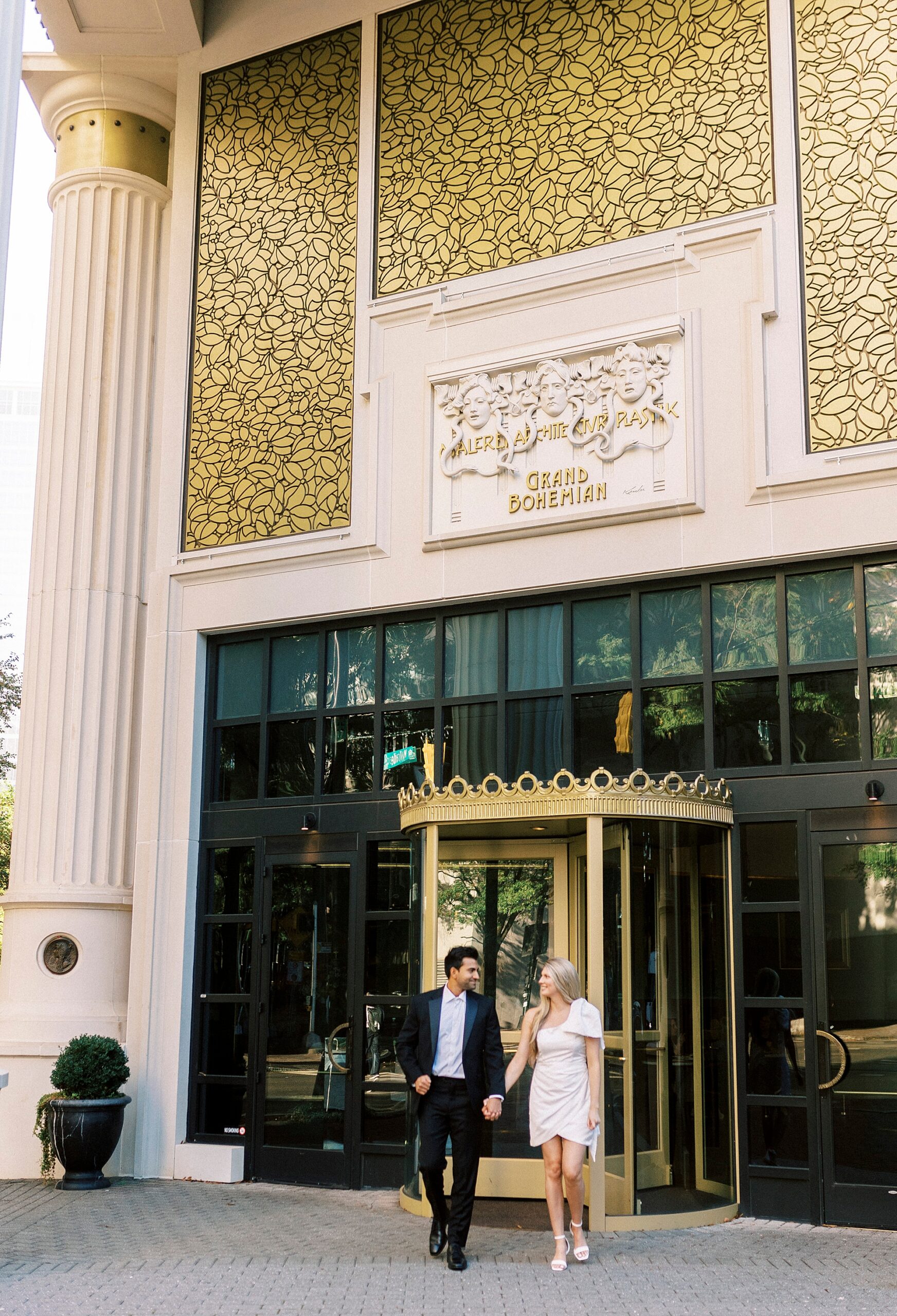 engaged couple walk outside gold doors of the Grand Bohemian Hotel in Charlotte NC