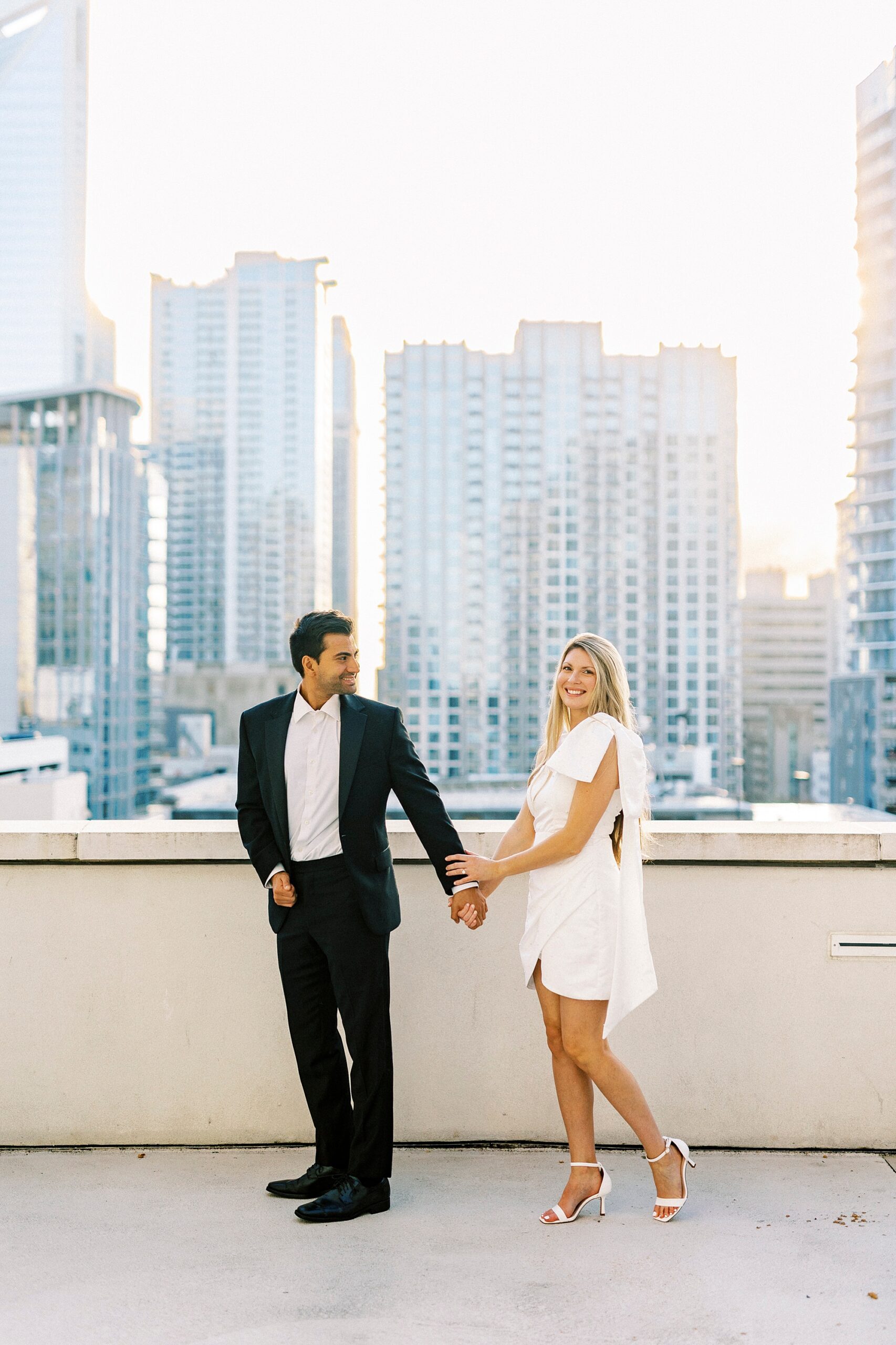couple holds hands walking in front of Charlotte NC skyline from the rooftop of the Grand Bohemian Hotel