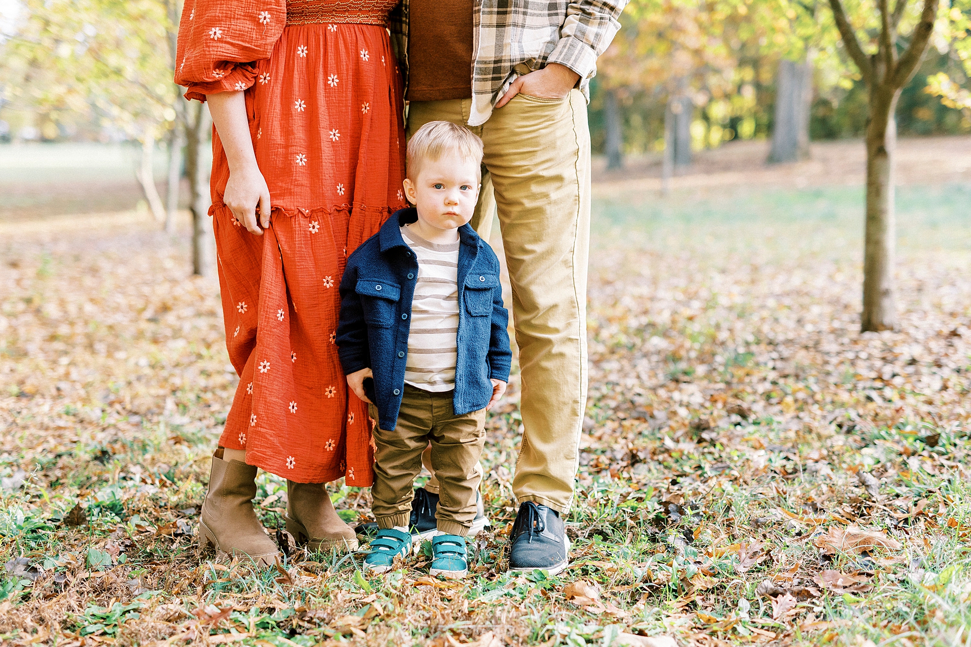 young boy stands between parents on fallen leaves in Rob Wallace Park