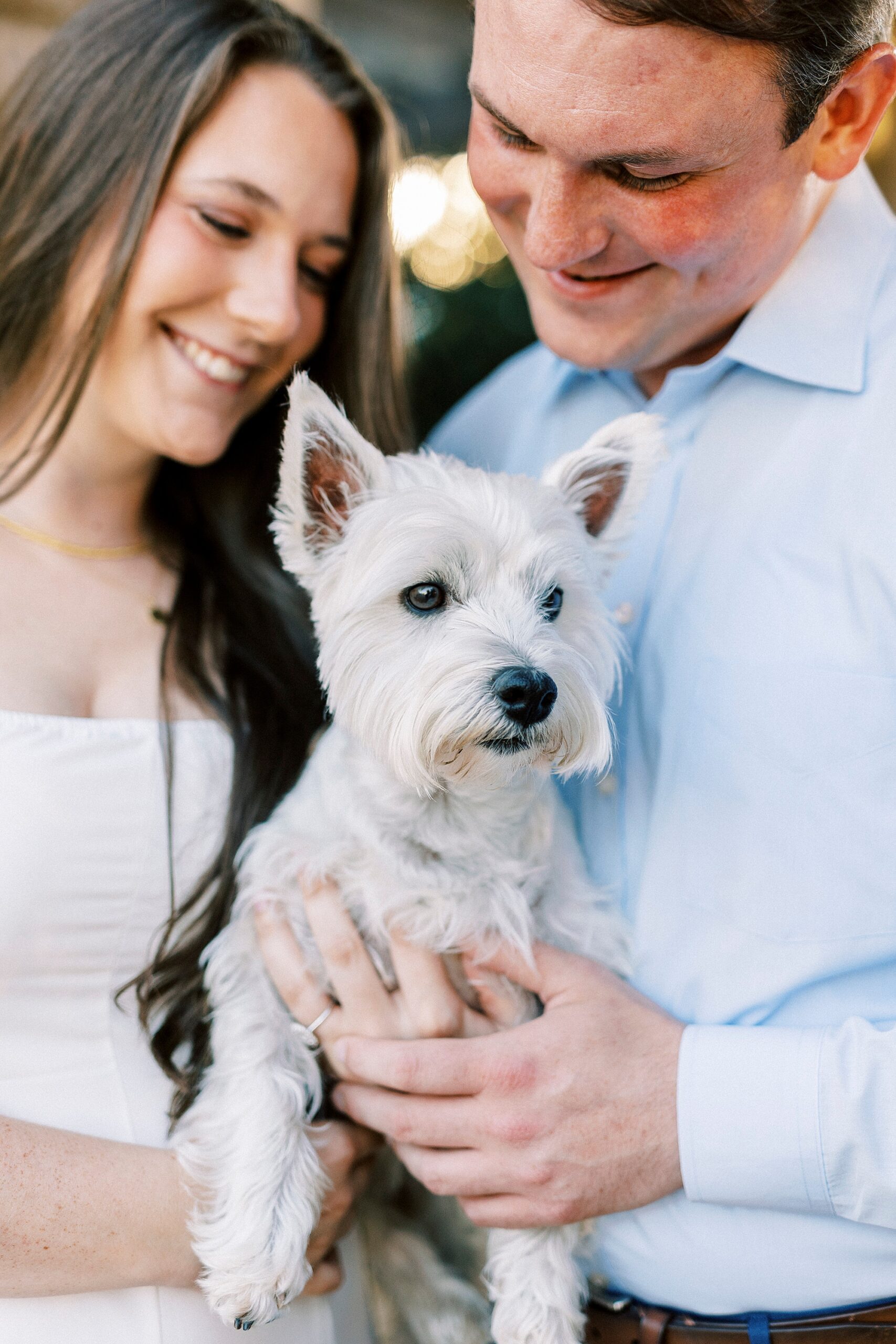 couple smiles at white dog between them during Midtown Park engagement photos 