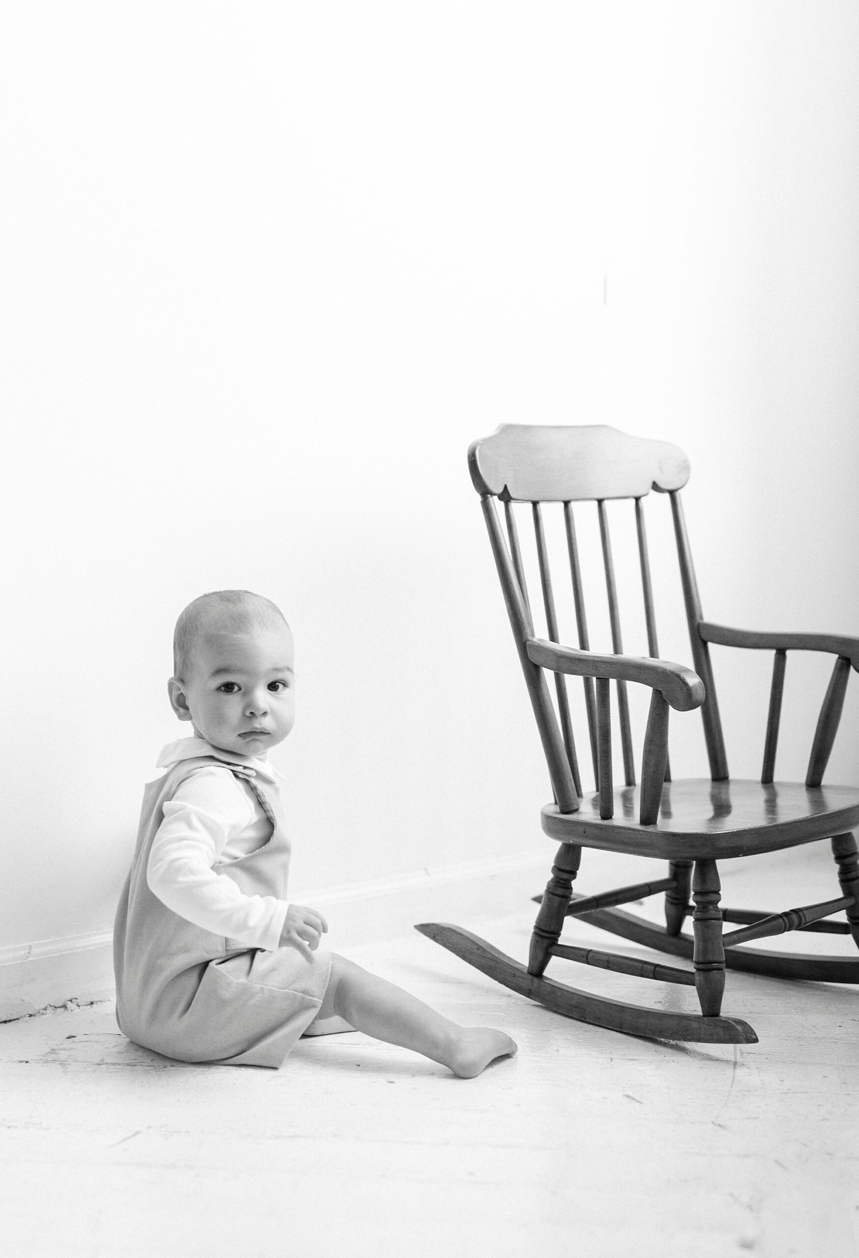 baby sits on floor by classic wooden rocking chair during first birthday photos in Concord NC studio