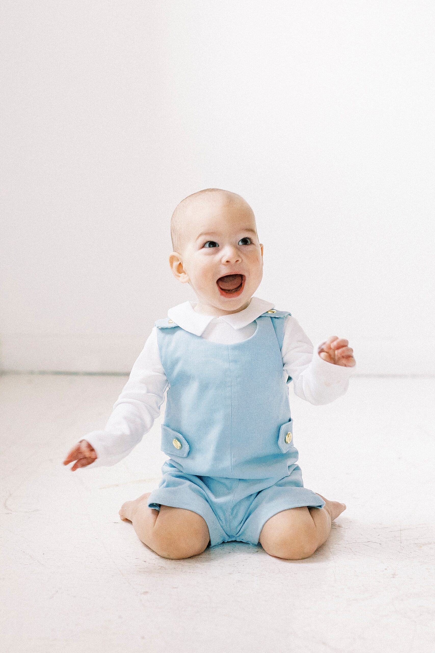 baby in blue romper laughs sitting in Concord NC studio for first birthday photos