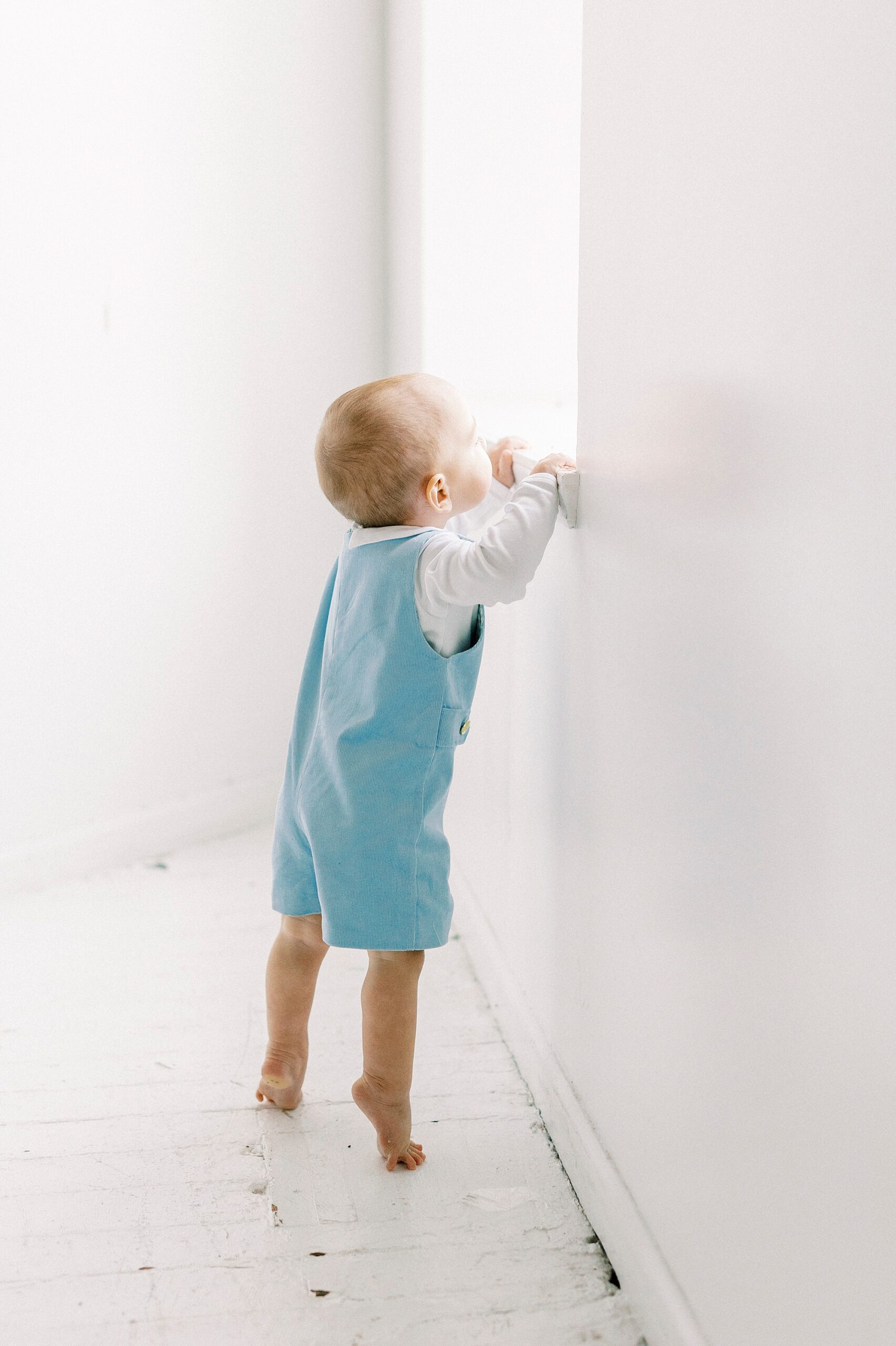 baby stands on tip toes to look out window during first birthday photos in Concord NC studio