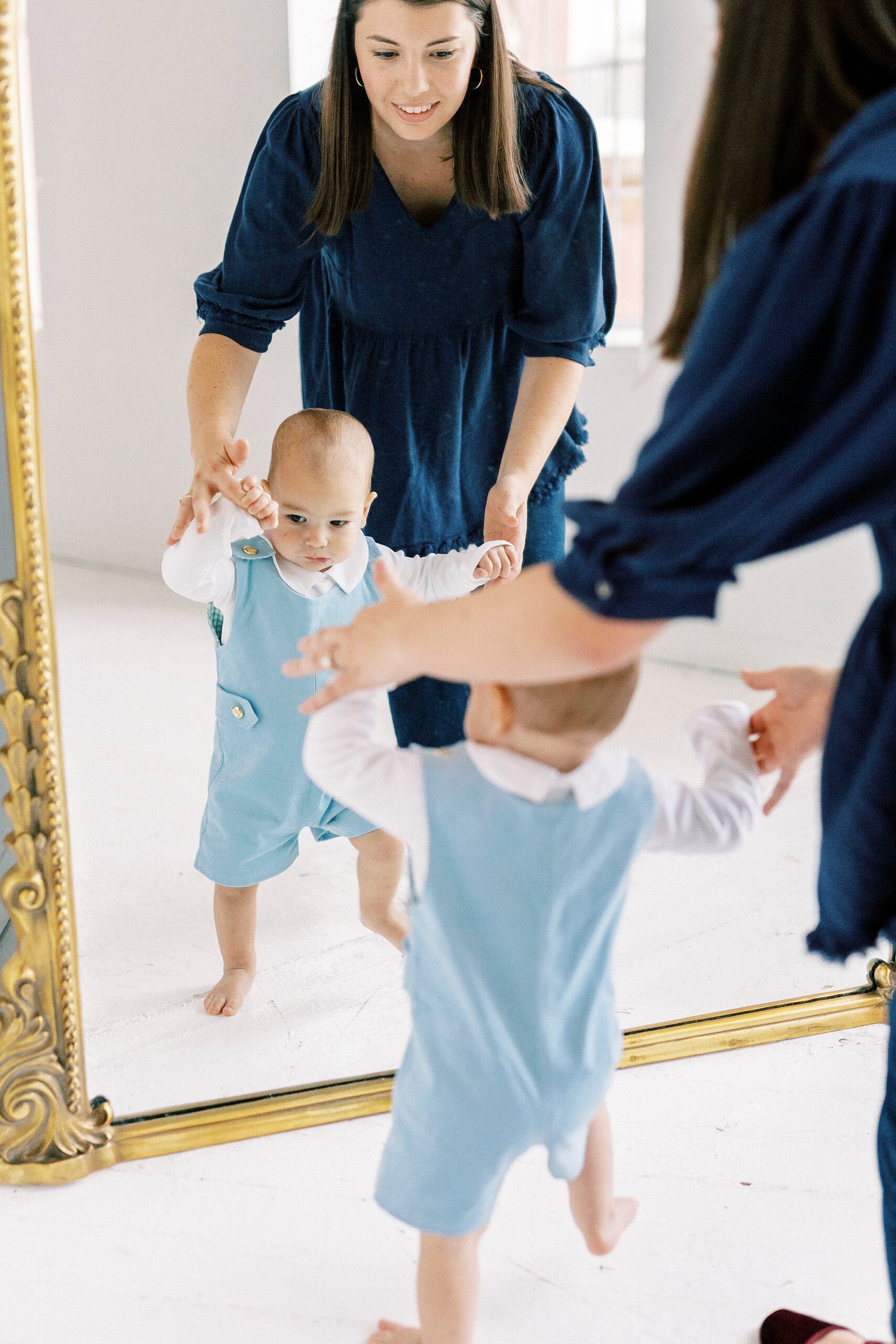 mom helps baby stand up in front of gold rimmed mirror during first birthday photos in Concord NC studio