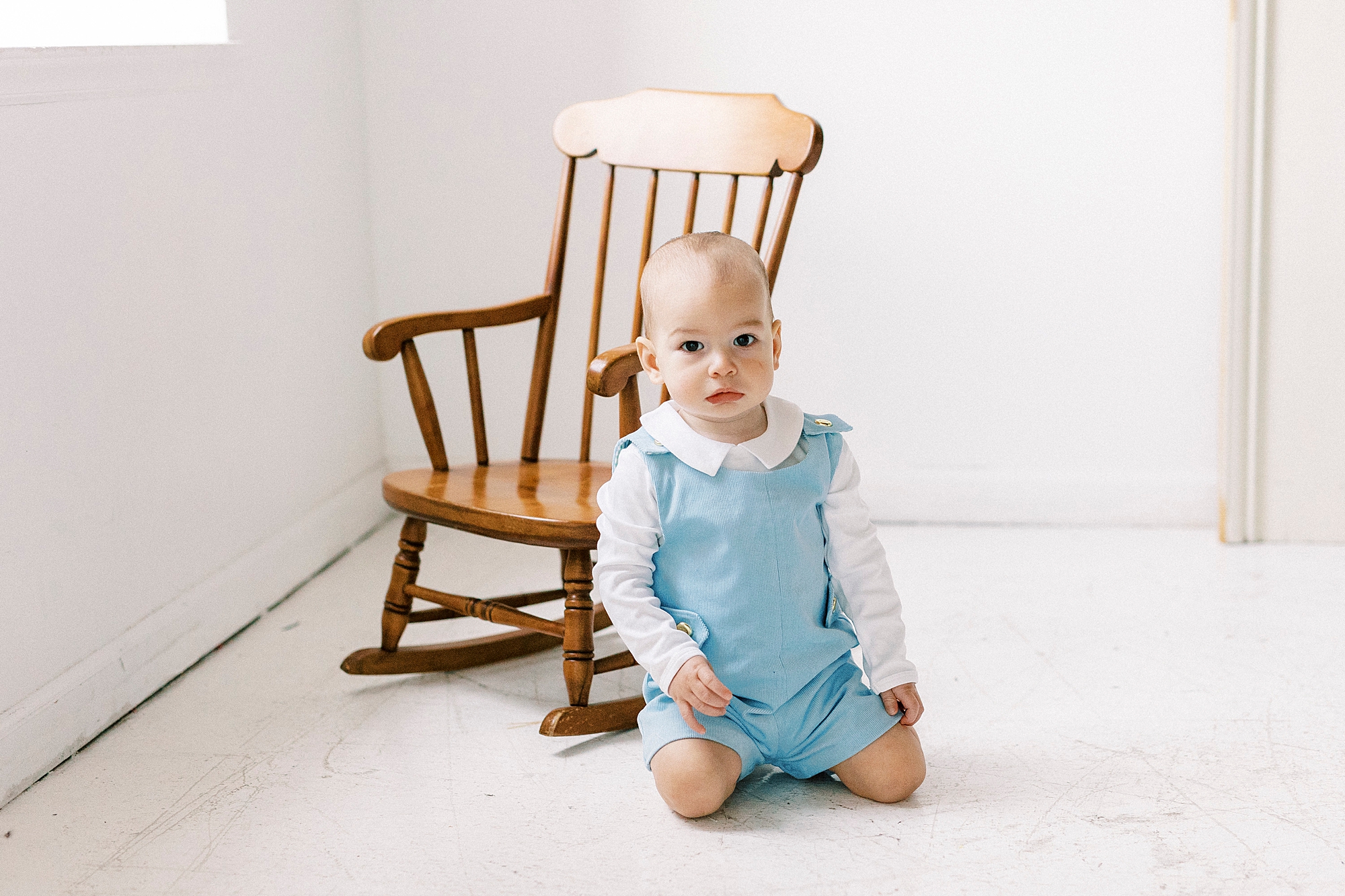 one year old sits in blue romper by wooden rocking chair during first birthday photo session
