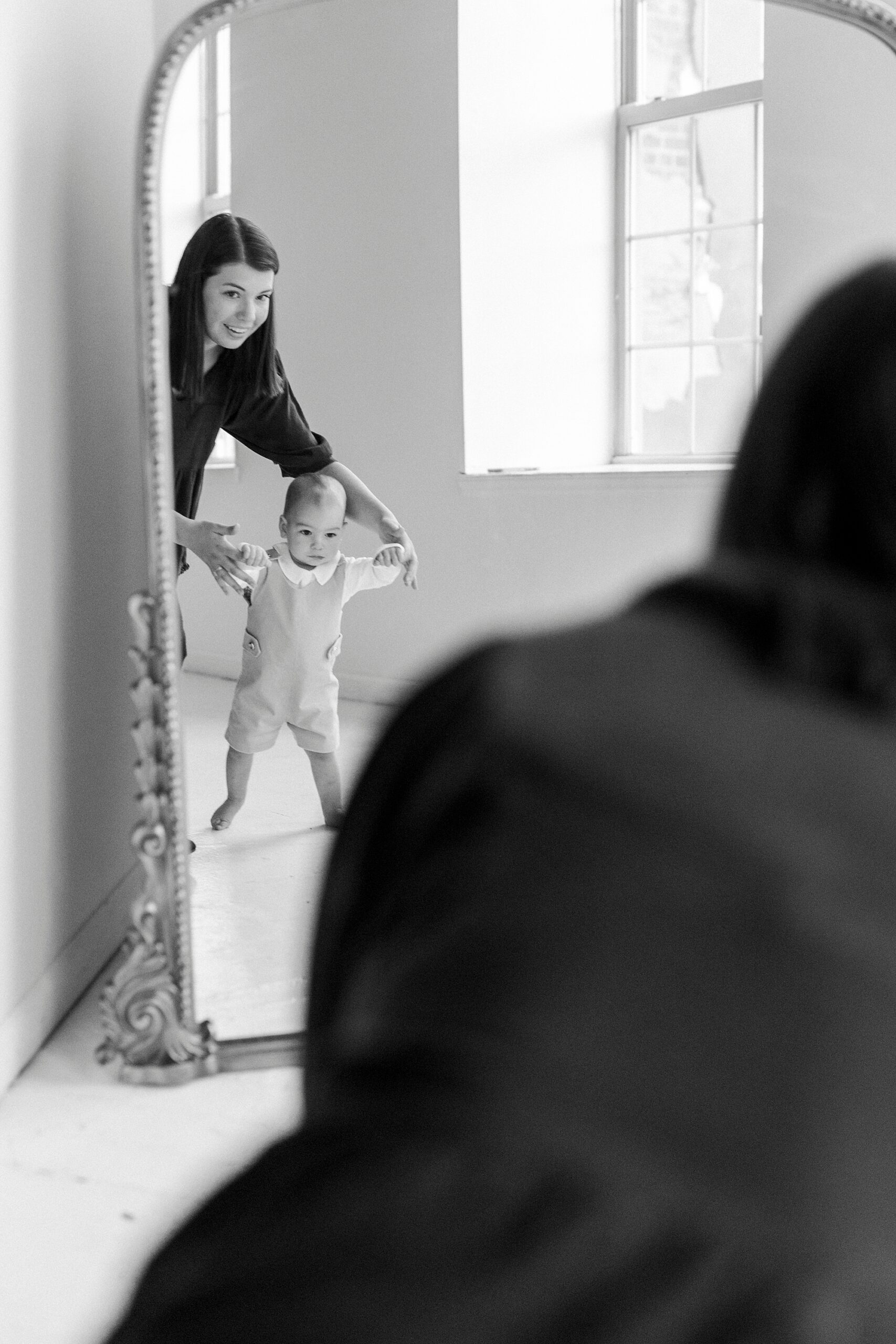 mom helps toddler stand in front of mirror inside Concord NC studio for first birthday photos