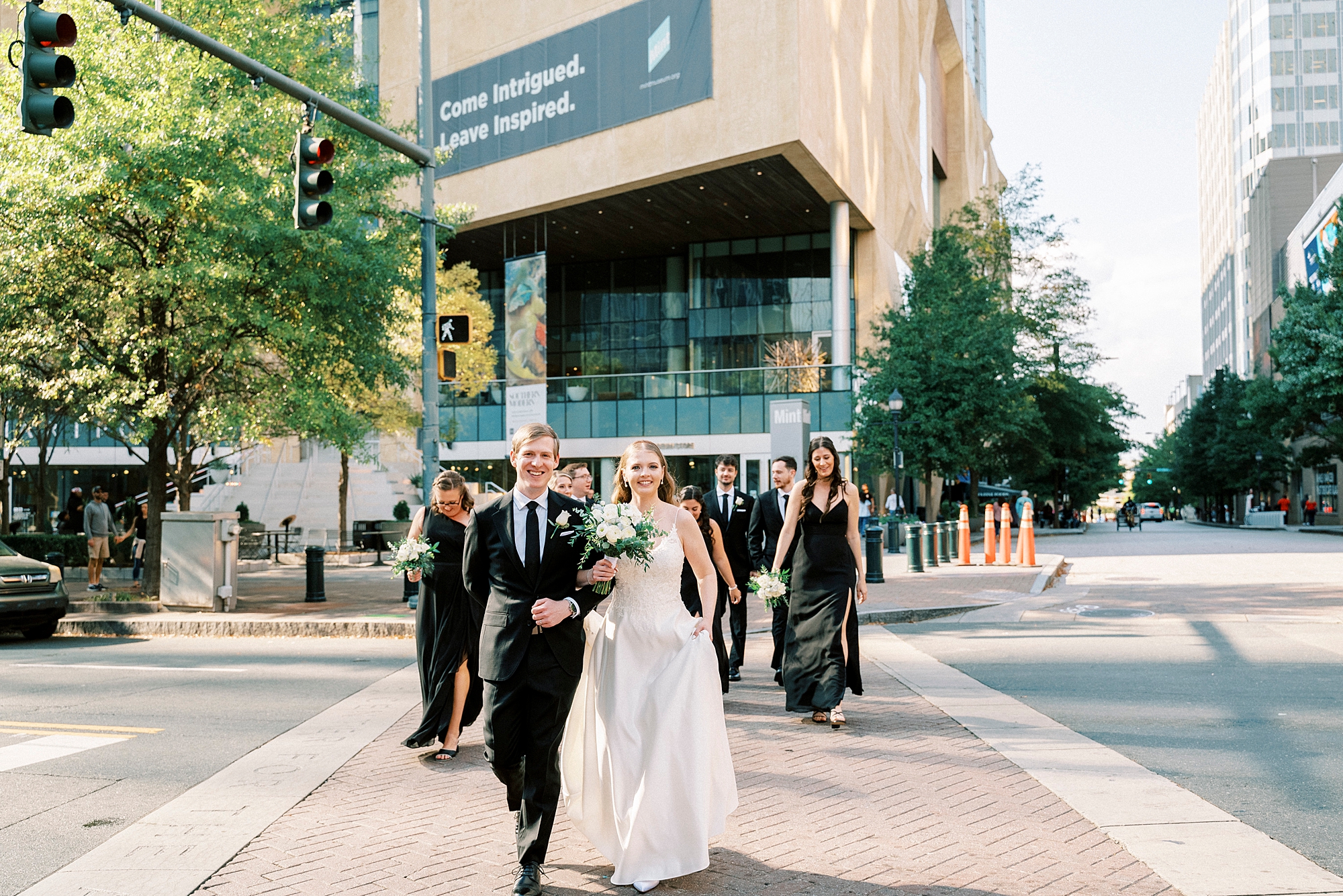 bride and groom walk with wedding party in black attire outside the Mint Museum Uptown