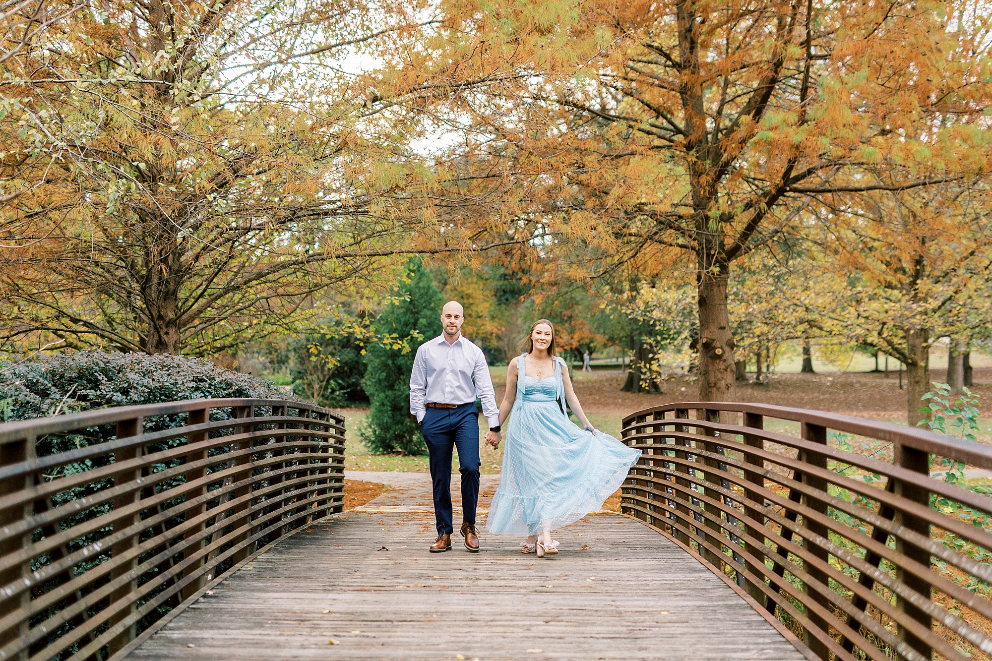 engaged couple holds hands walking across wooden bridge inside Glencairn Garden