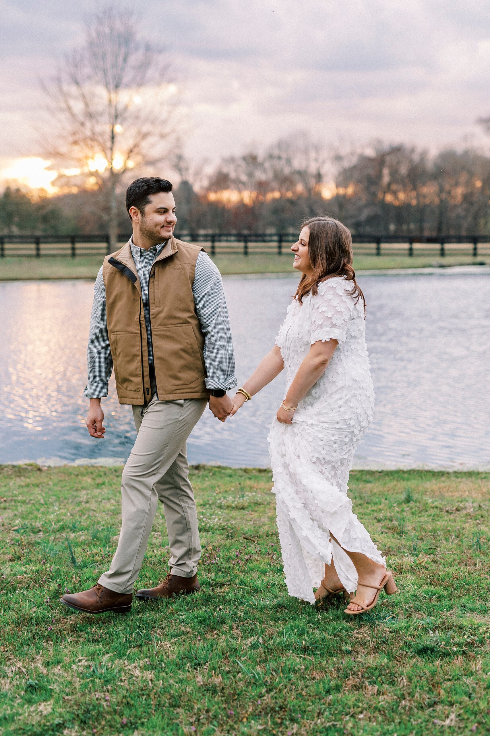 couple holds hands walking along pond during engagement session at Morning Glory Farm