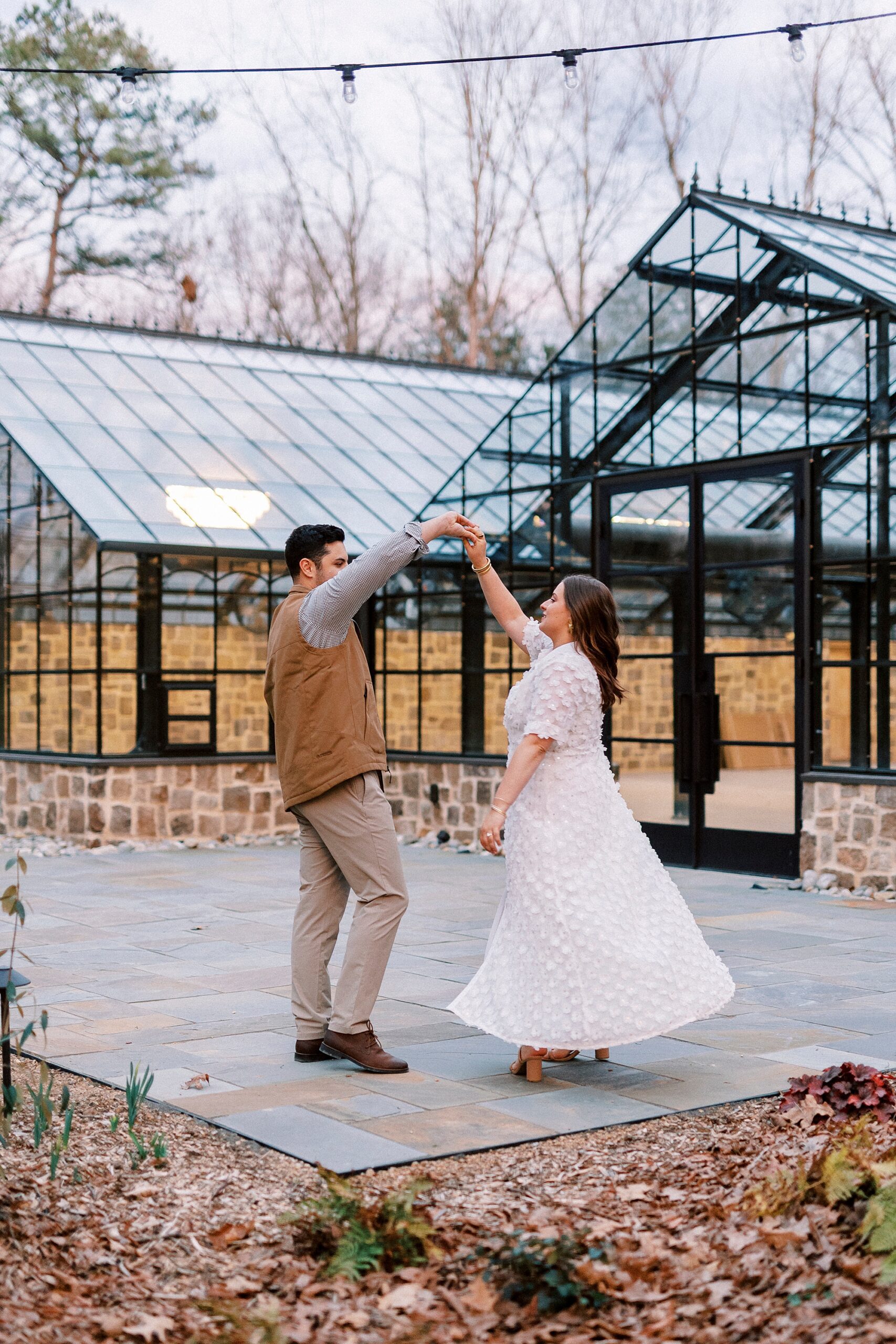 man twirls woman in front of greenhouse during engagement session at Morning Glory Farm