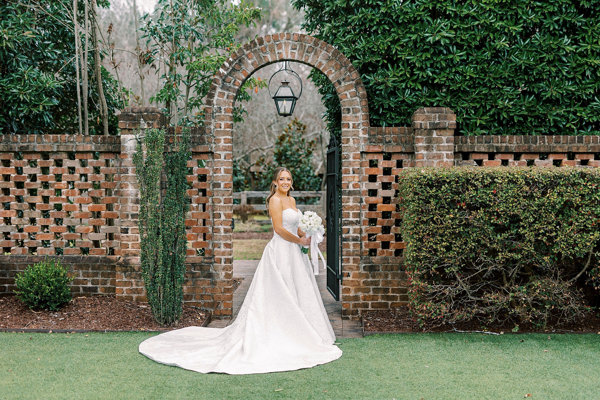 bride holds bouquet of white flowers in front of brick archway in gardens at the Sutherland in Wake Forest NC