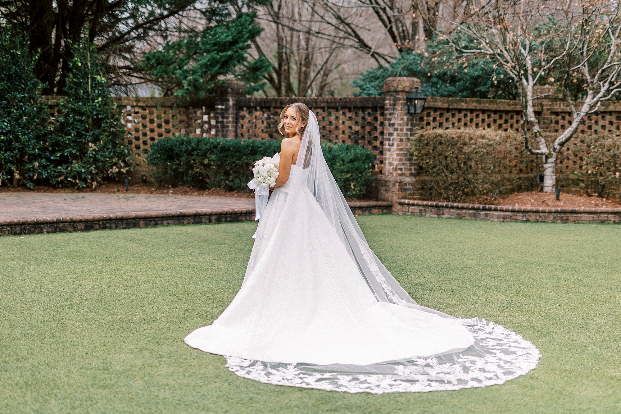 bride turns looking over shoulder holding bouquet of white flowers in courtyard at the Sutherland in Wake Forest NC