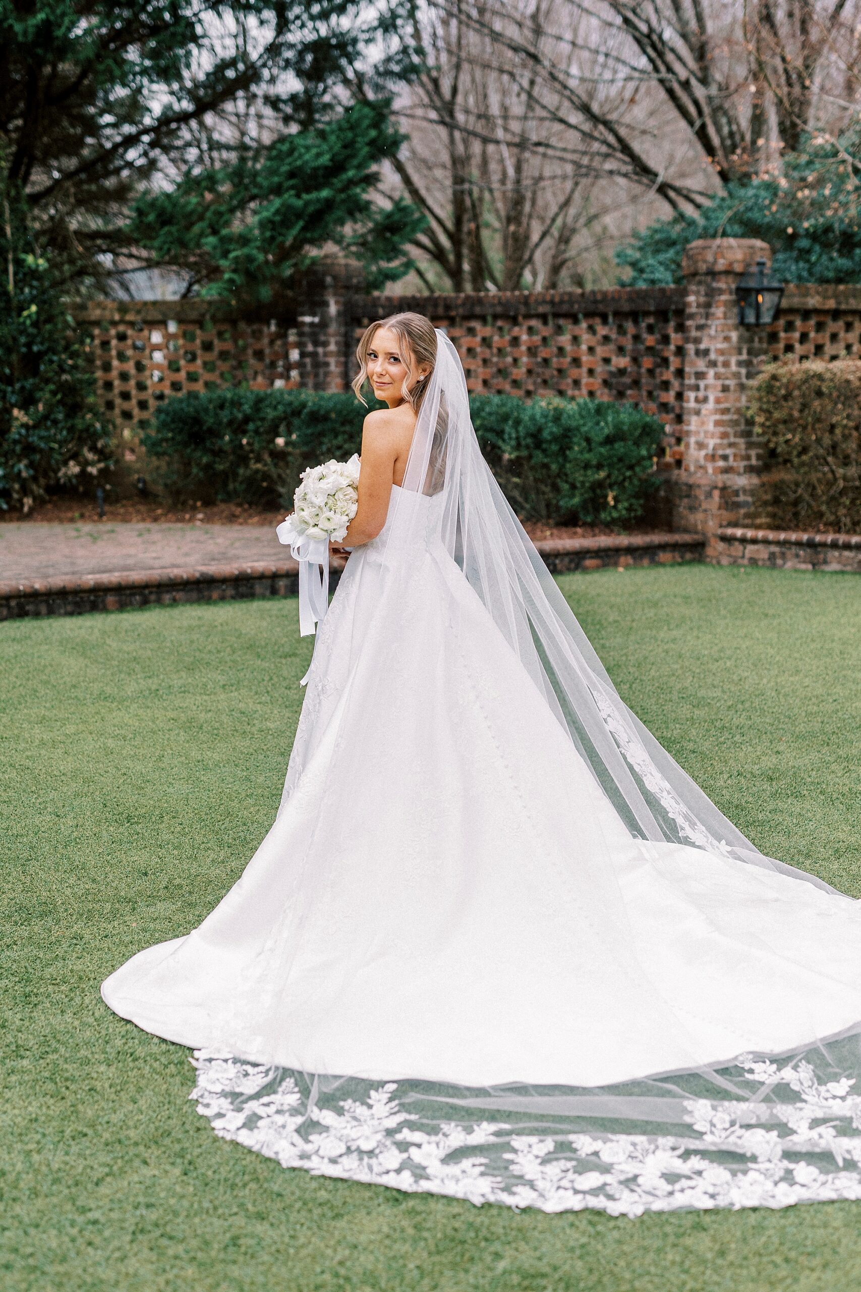 bride stands on lawn at bridal portraits at the Sutherland with veil pulled behind him