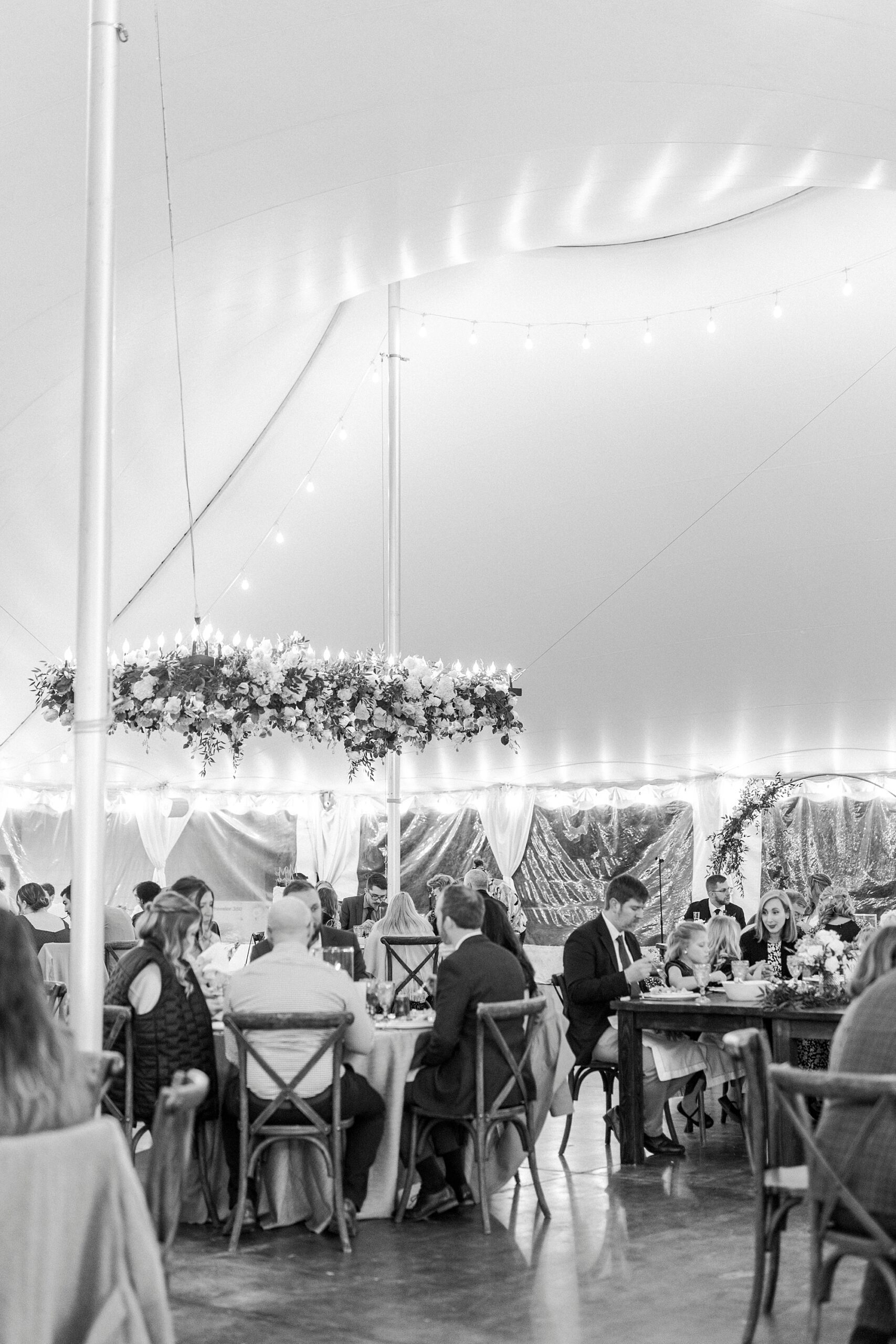 wedding guests sit talking during dinner under hanging installation in tent at Summerfield Farms