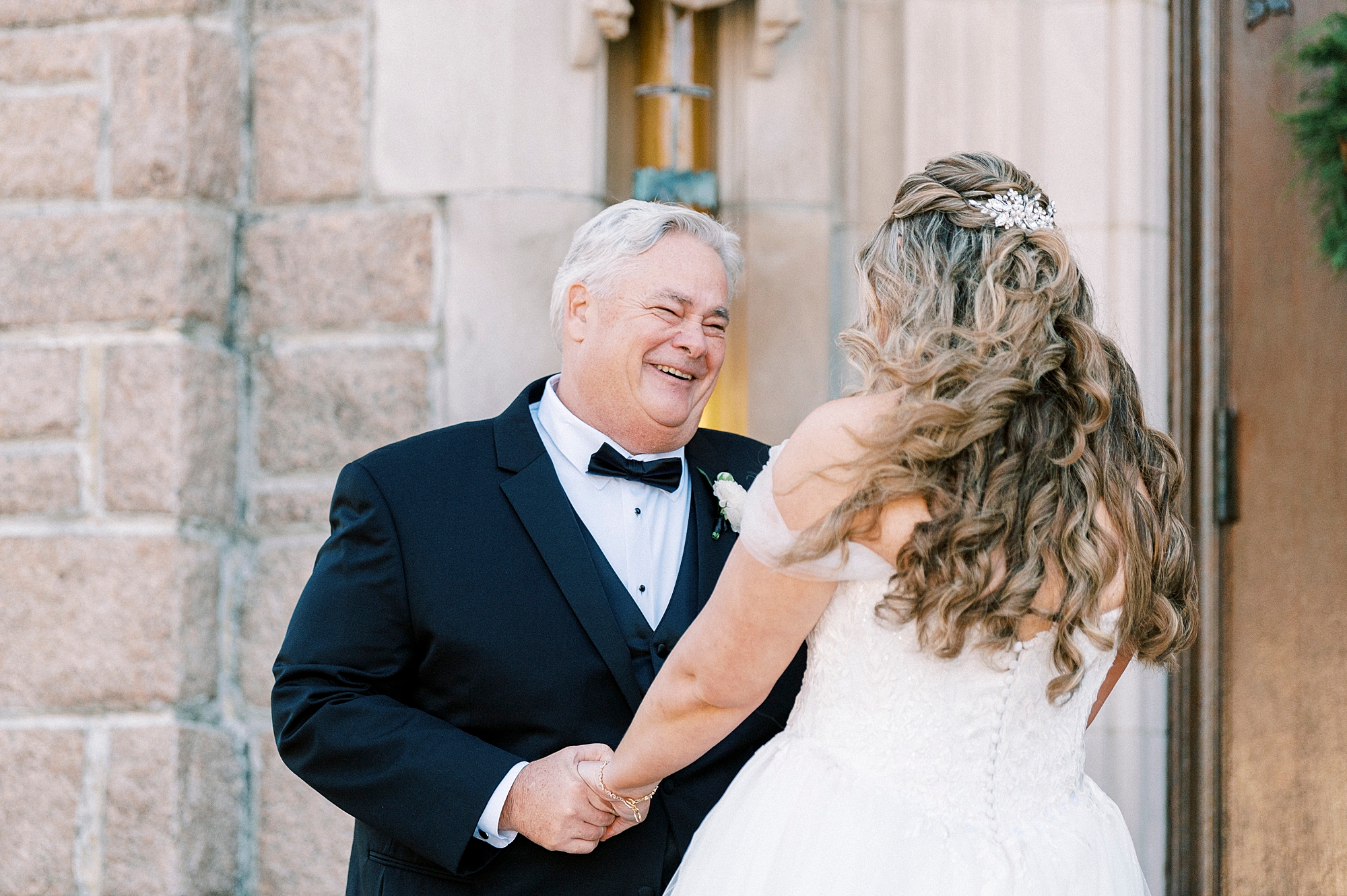 dad holds bride's hands and looks at her smiling during first look outside Our Lady of Grace
