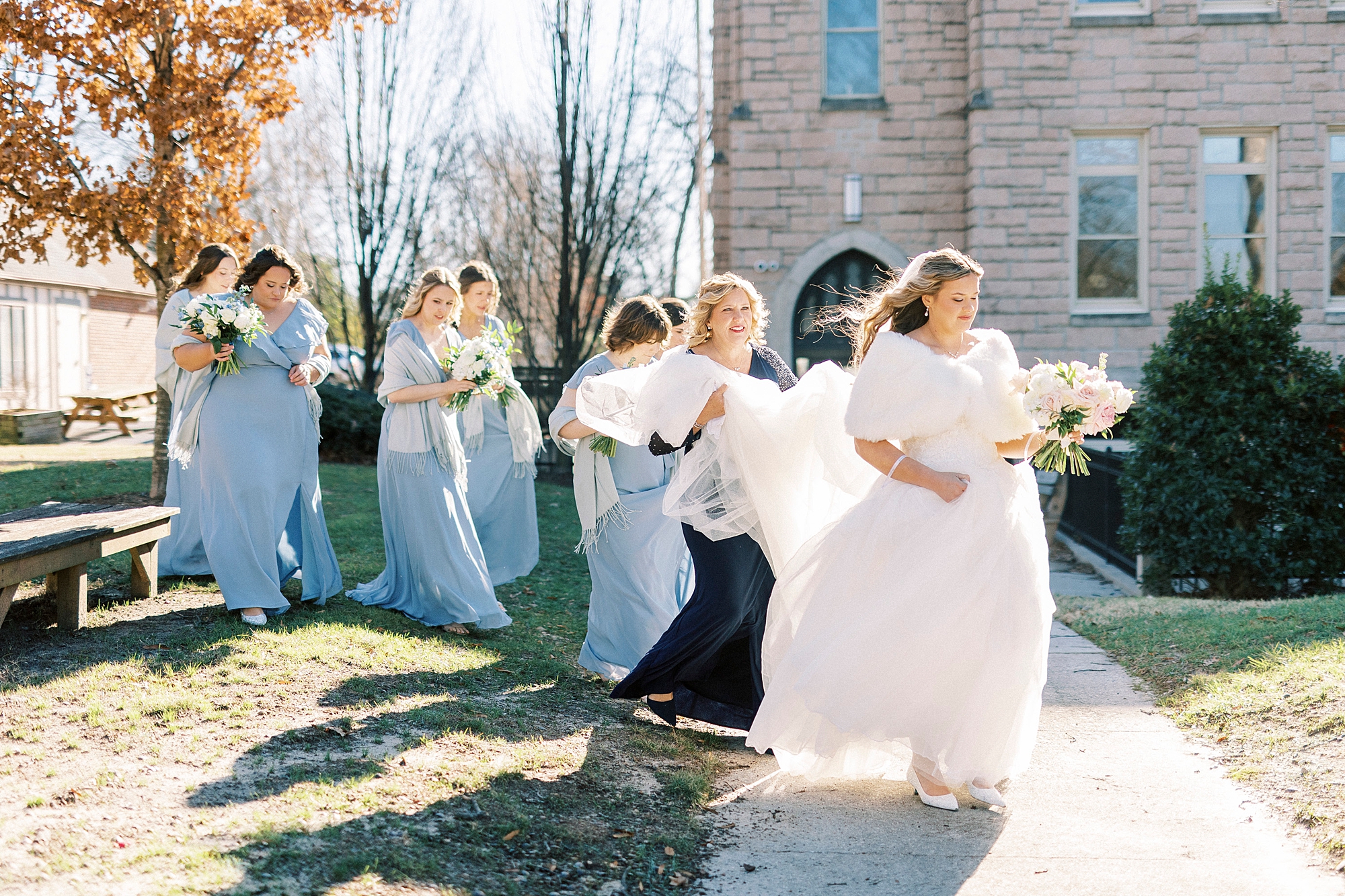 bridesmaids in light bleu gowns carry bride's veil and dress outside Our Lady of Grace
