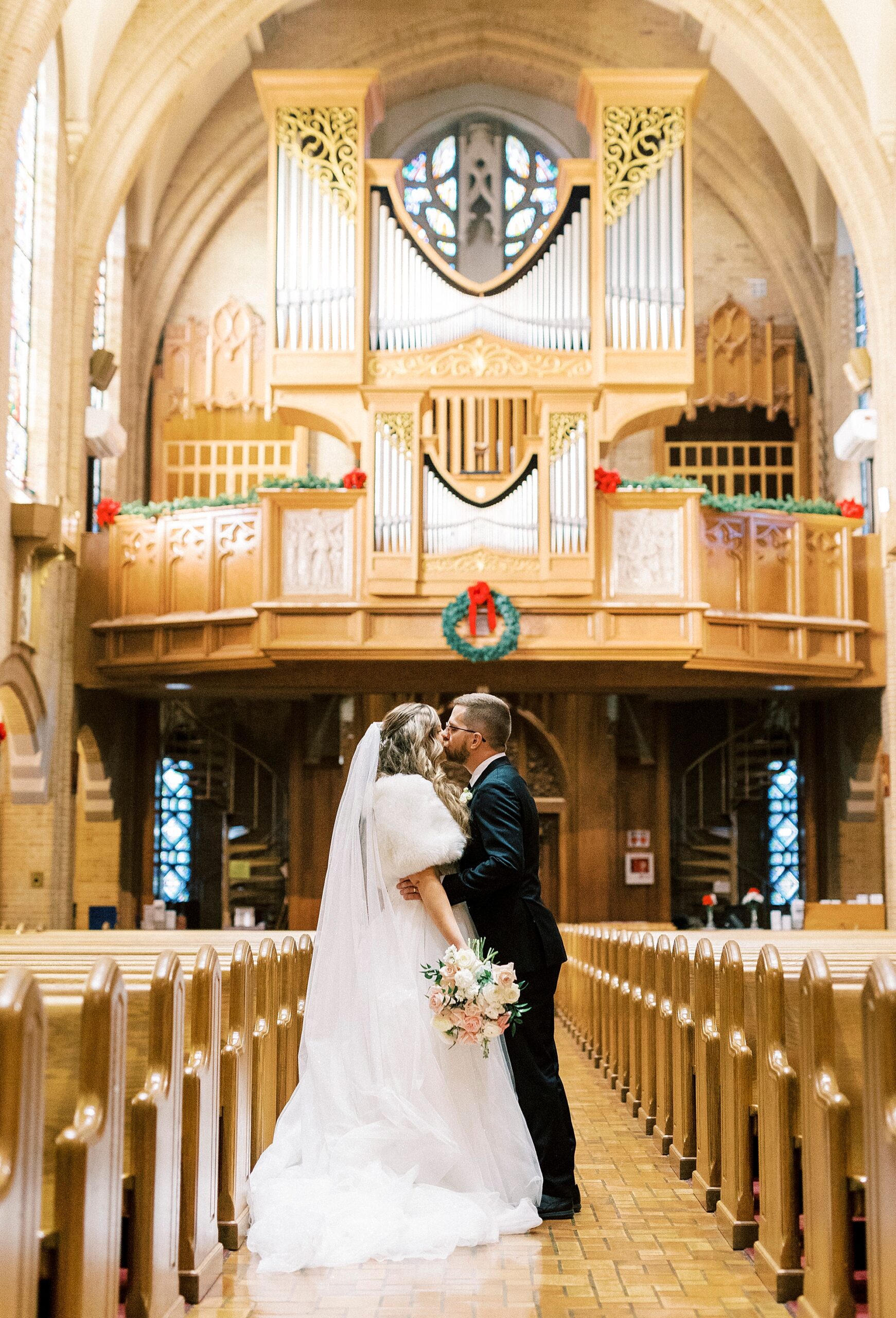 bride and groom kiss in aisle under balcony with holiday wreaths hanging at Our Lady of Grace