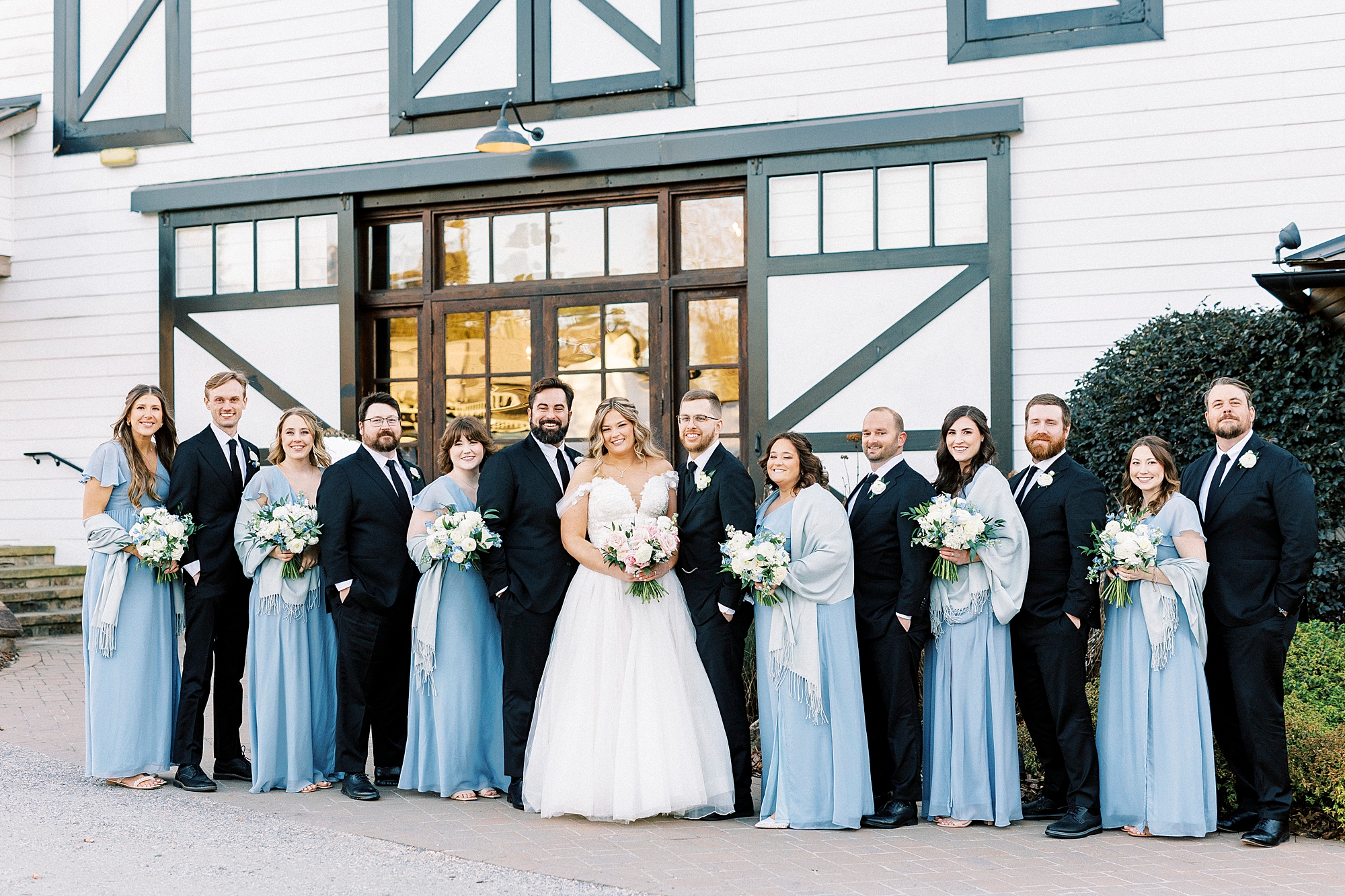 bride and groom stand outside Summerfield Farms with wedding party in light blue and black attire