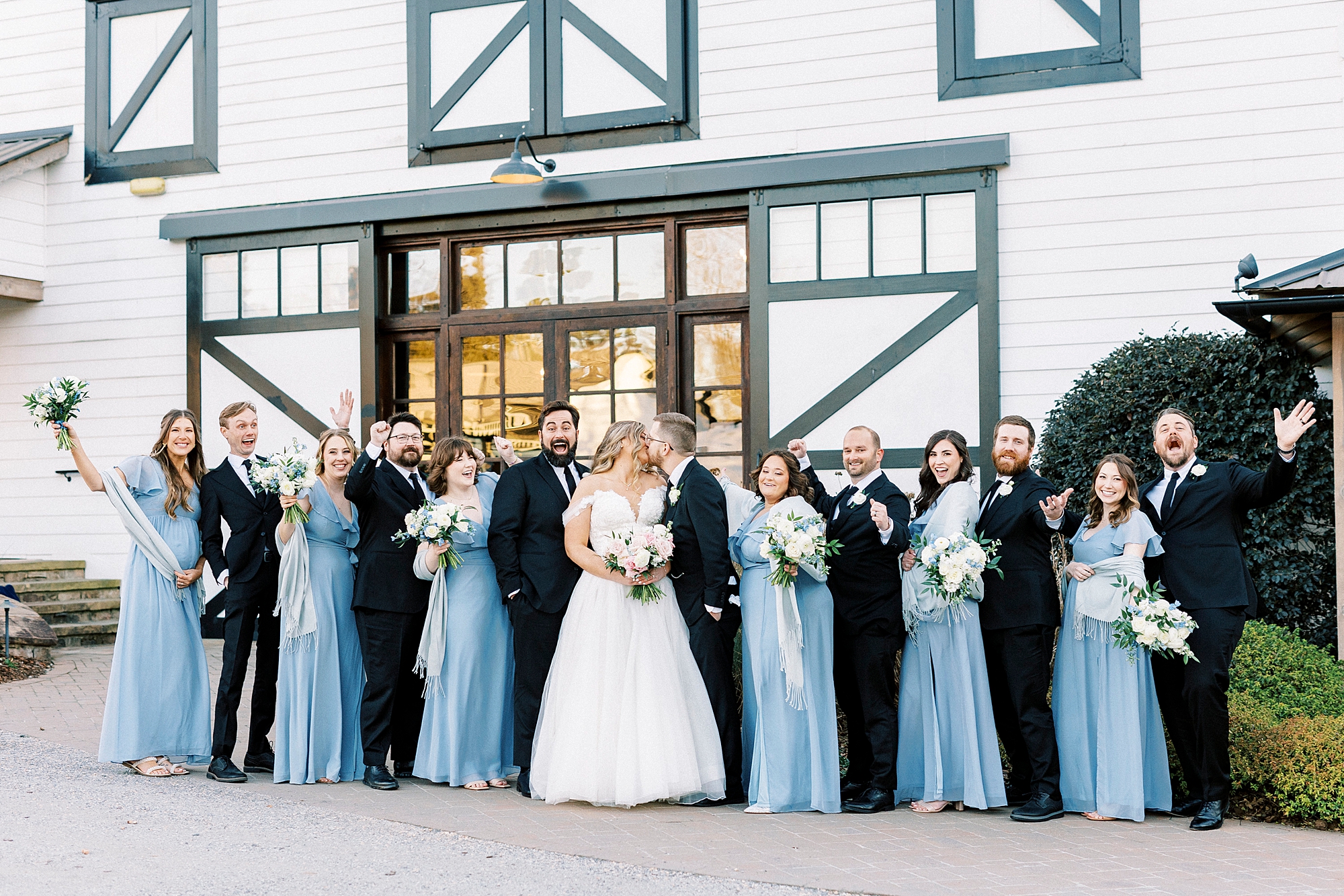 bride and groom kiss with wedding party standing outside barn at Summerfield Farms