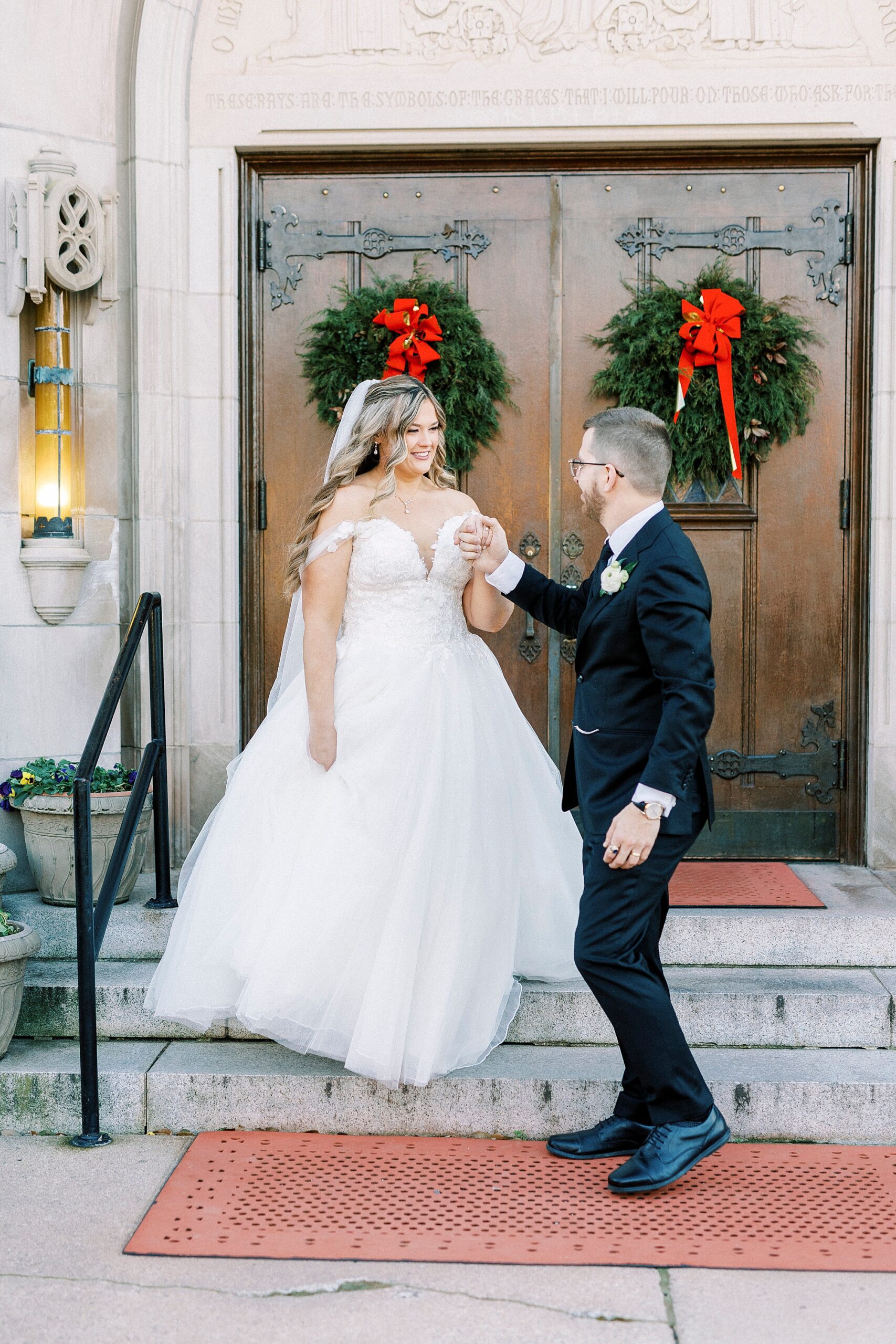 groom leads bride down steps outside Our Lady of Grace with wreaths on wooden doors 