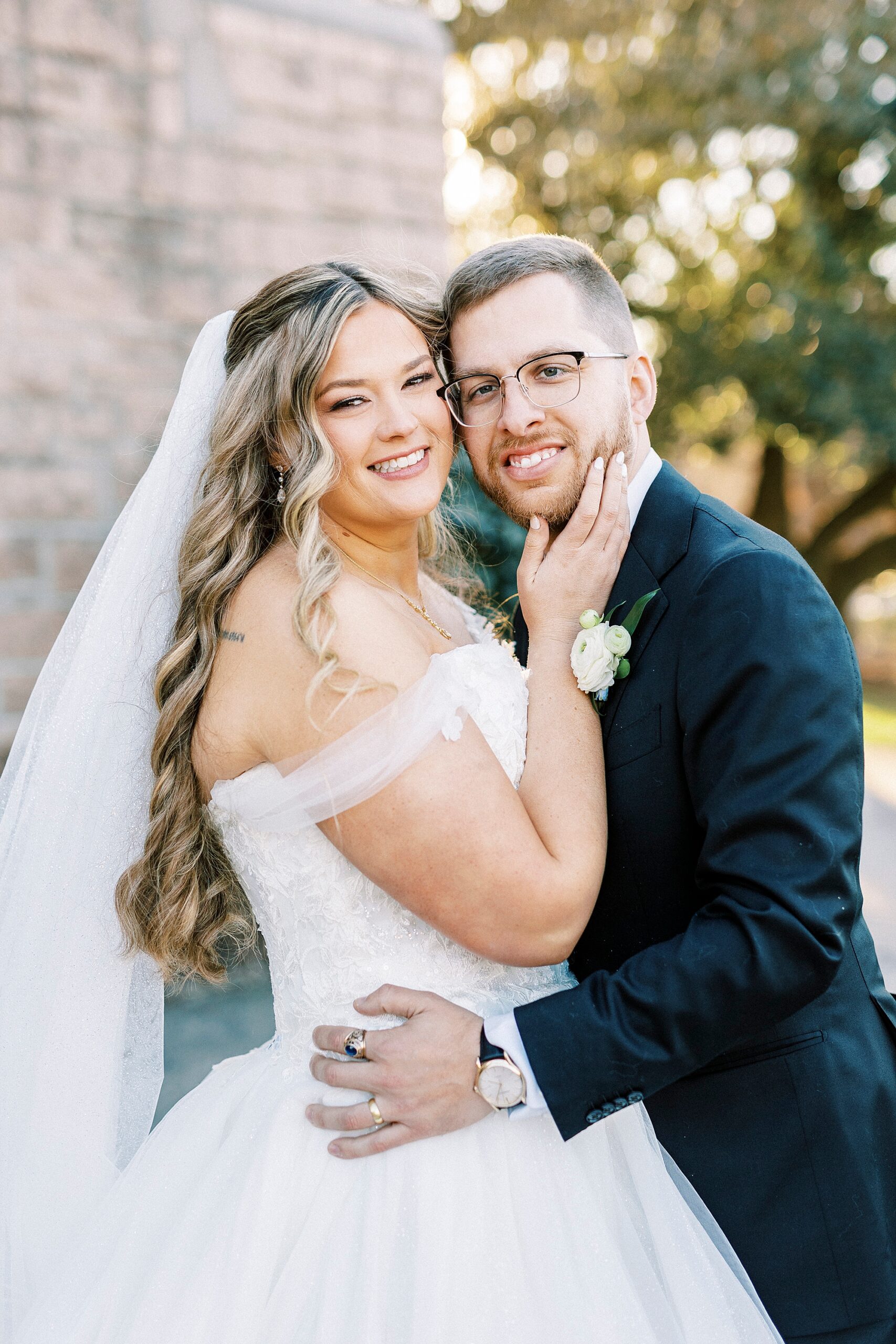 bride hugs groom holding his chin outside stone walls of Our Lady of Grace