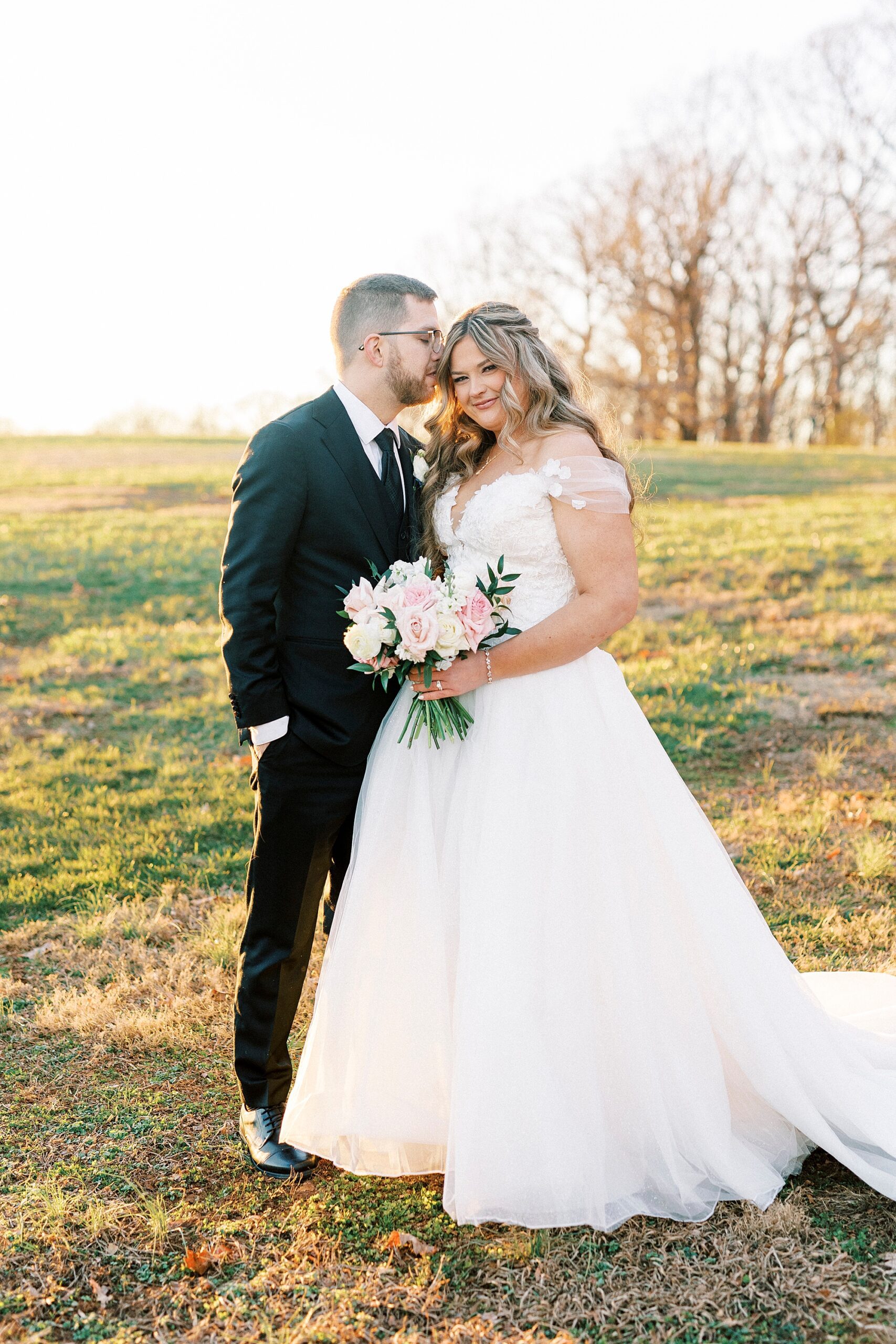 groom hugs bride to him kissing her cheek on lawn at Summerfield Farms