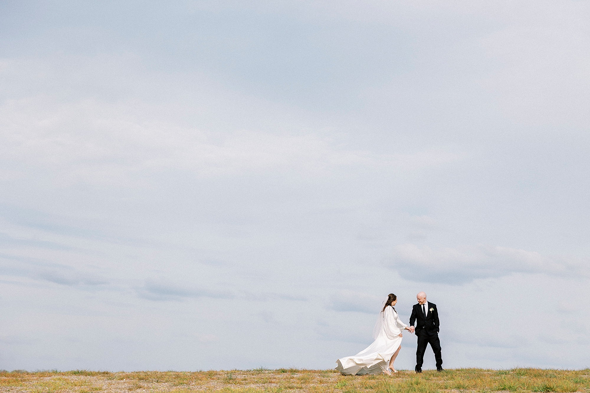 bride and groom hold hands in front of blue sky on hill at Sixty Acres Farm