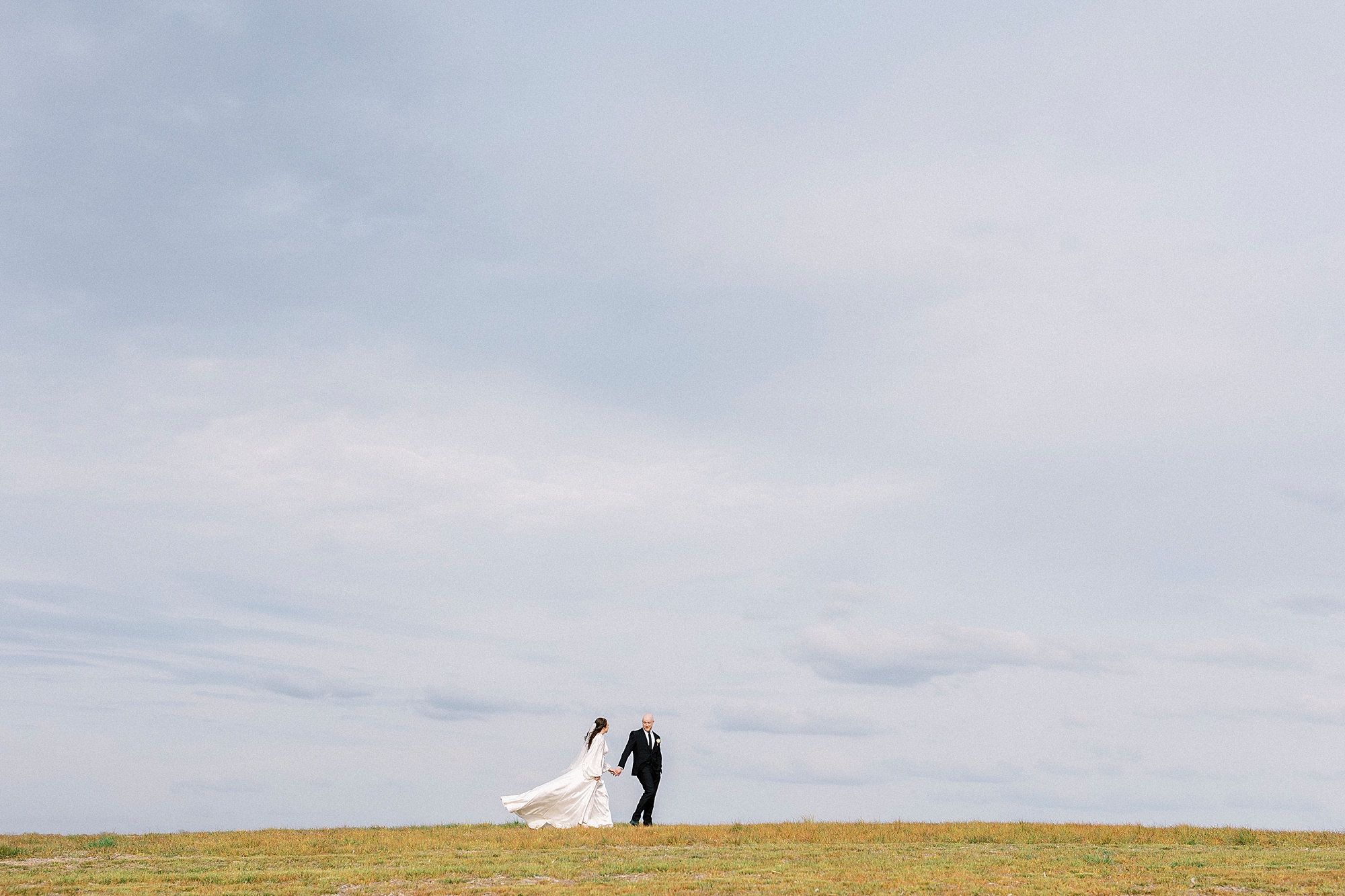 bride and groom hold hands in front of blue sky on hill at Sixty Acres Farm