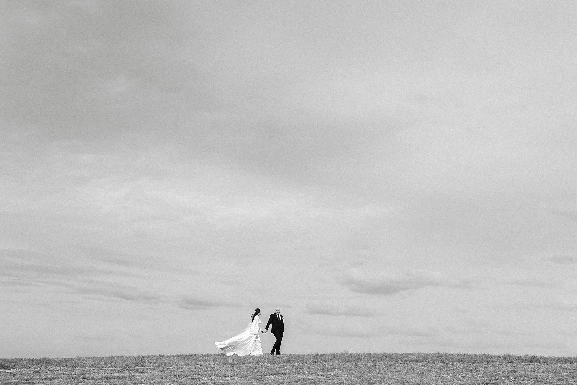 bride and groom hold hands in front of blue sky on hill at Sixty Acres Farm