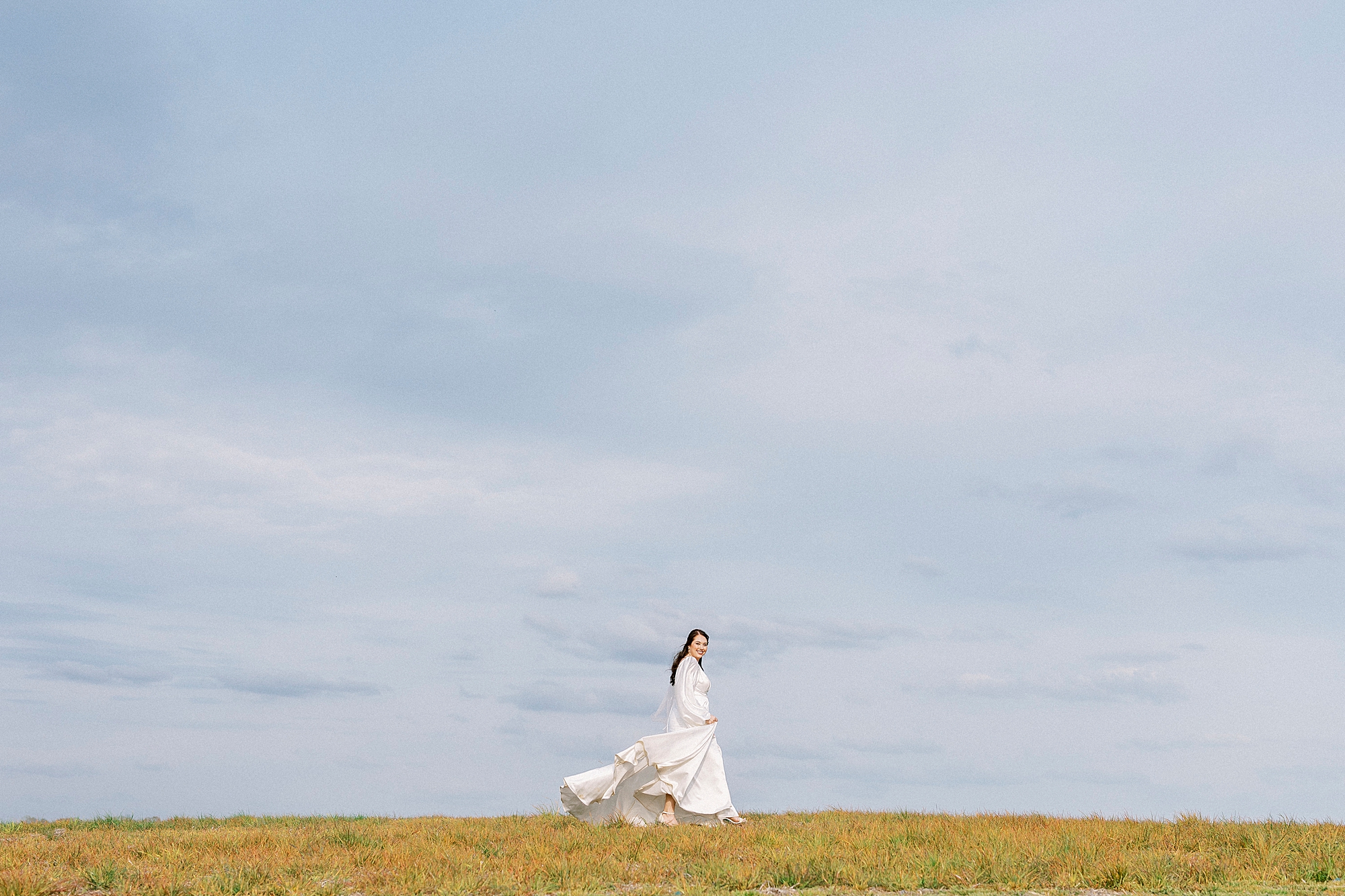 bride poses in wedding gown in front of blue sky on hill at Sixty Acres Farm