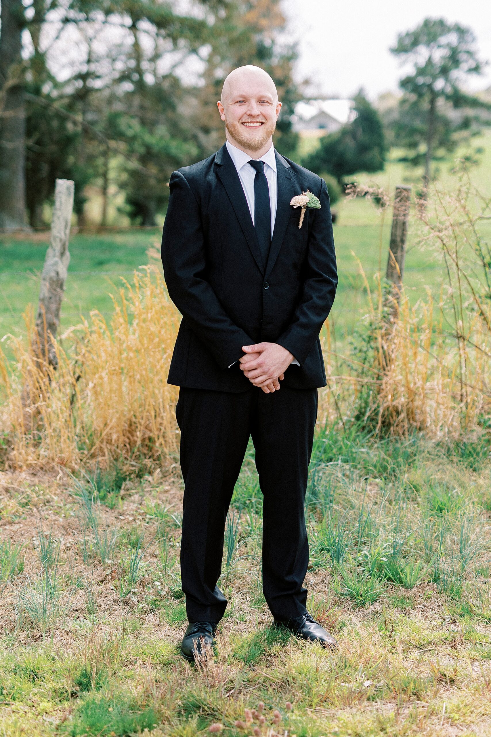 groom stands in black suit on edge of pasture at Sixty Acres Farm
