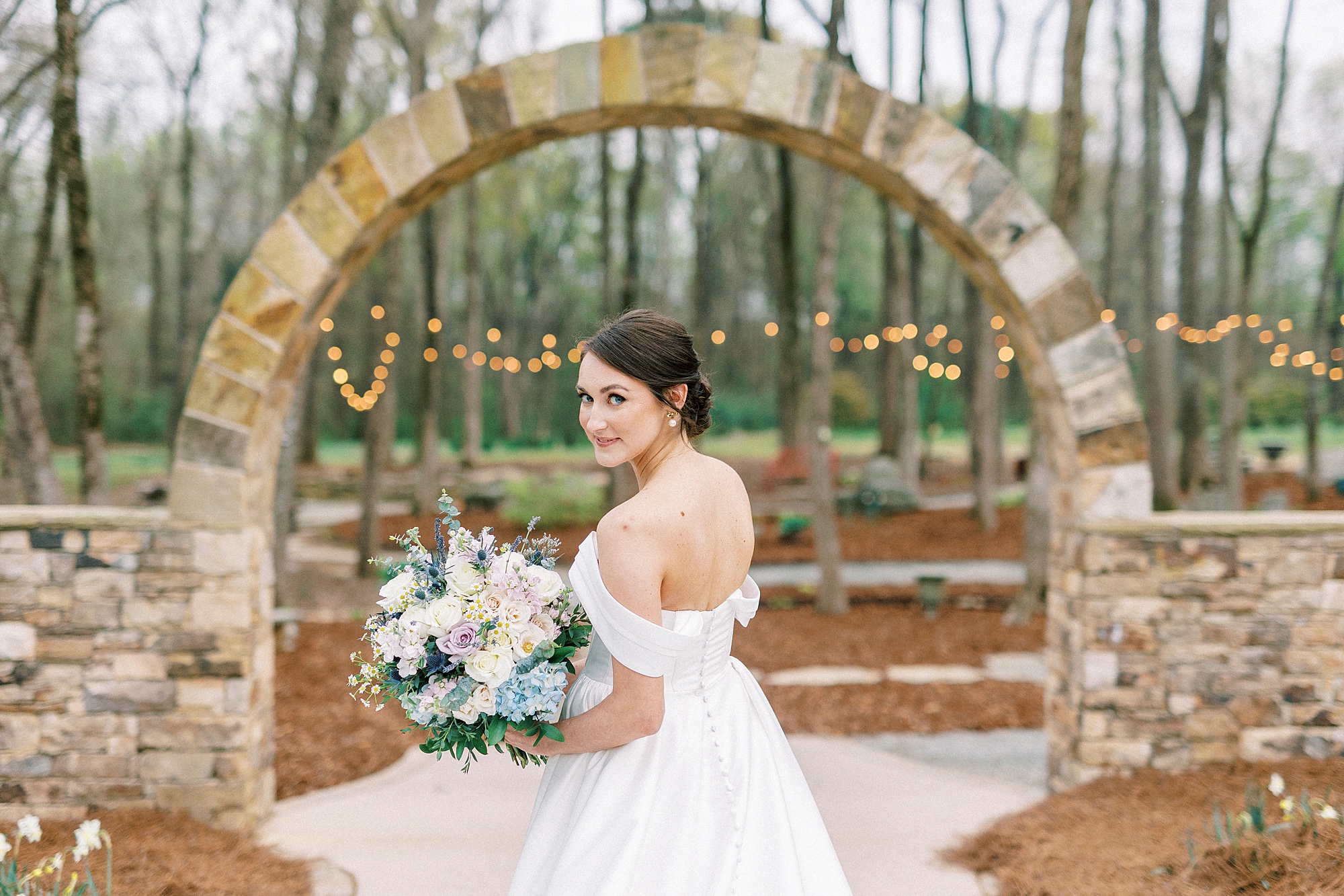 bride holds bouquet of spring flowers by stone arch in gardens of Low Meadows Estate