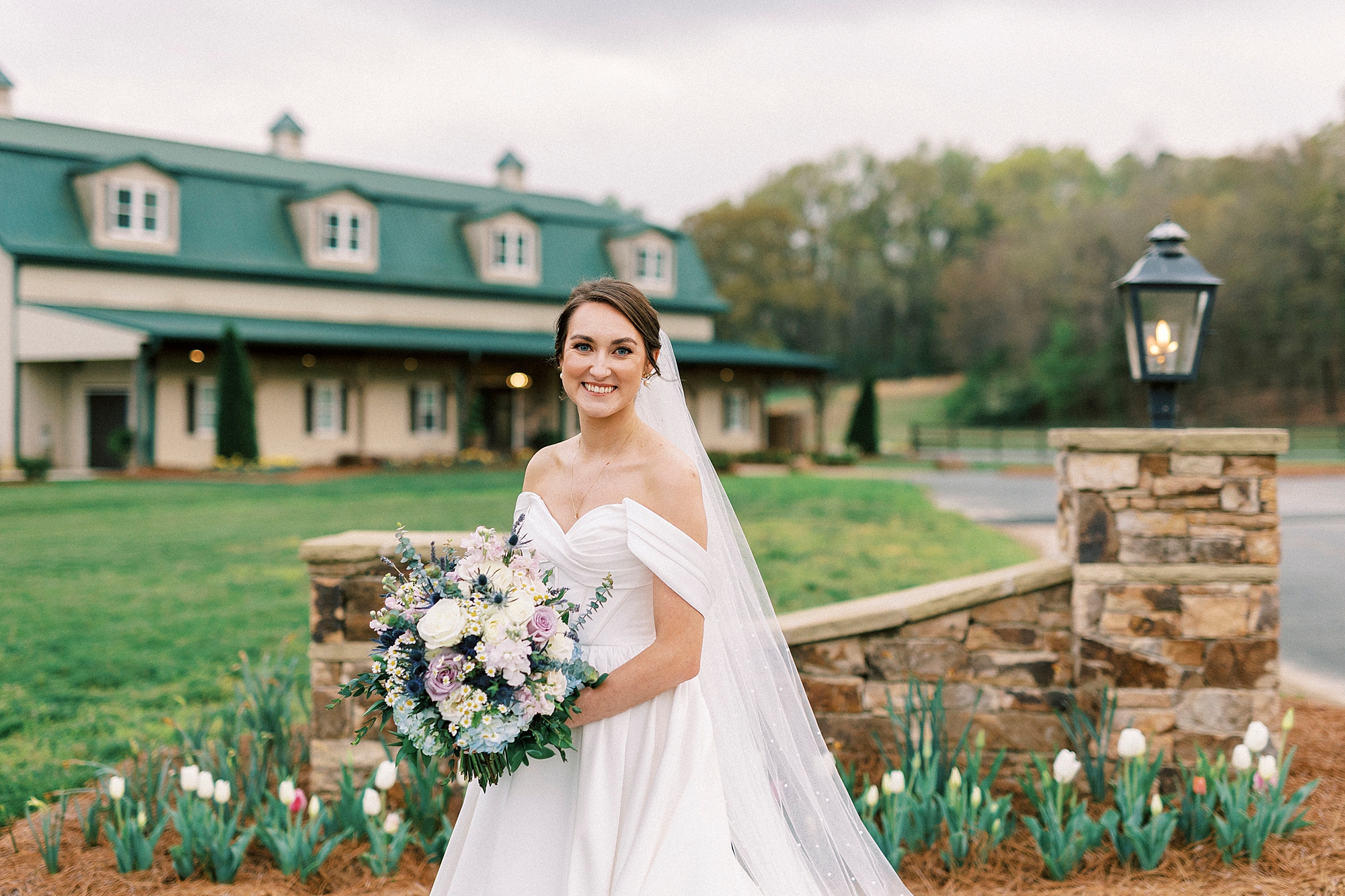 bride stands by stone gate and farmhouse at Low Meadows Estate