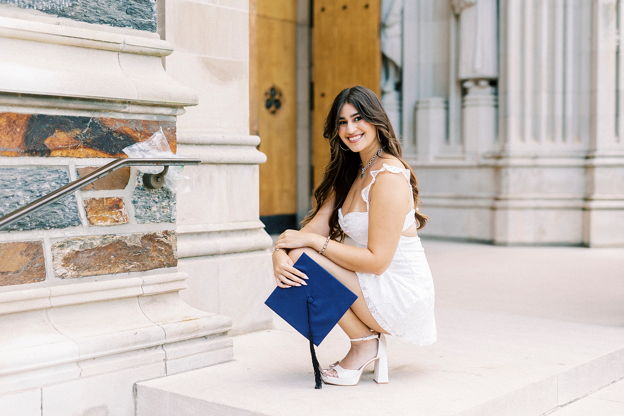 senior kneels down in white dress with blue graduation cap
