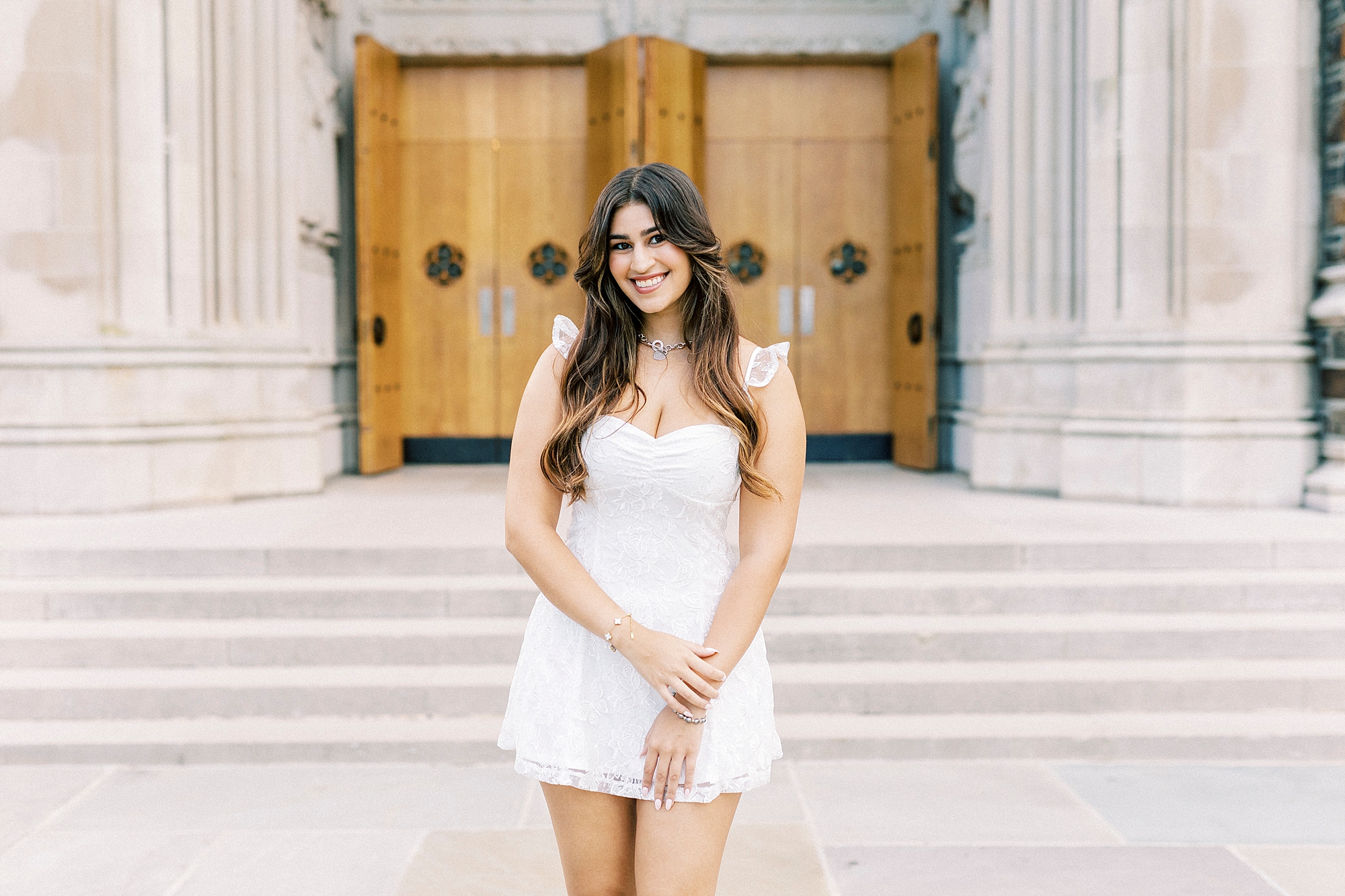 college senior poses in white dress outside steps and wooden of Duke Chapel