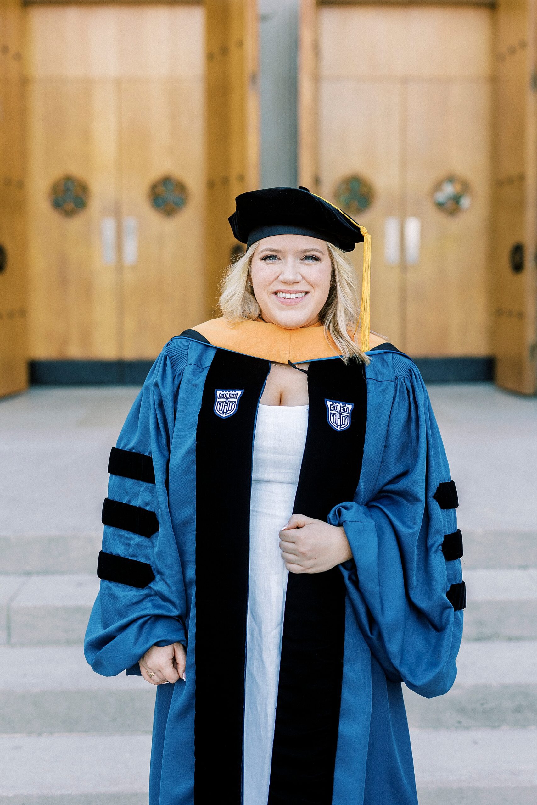 woman in blue and black gown poses outside wooden doors during graduation portraits at Duke University