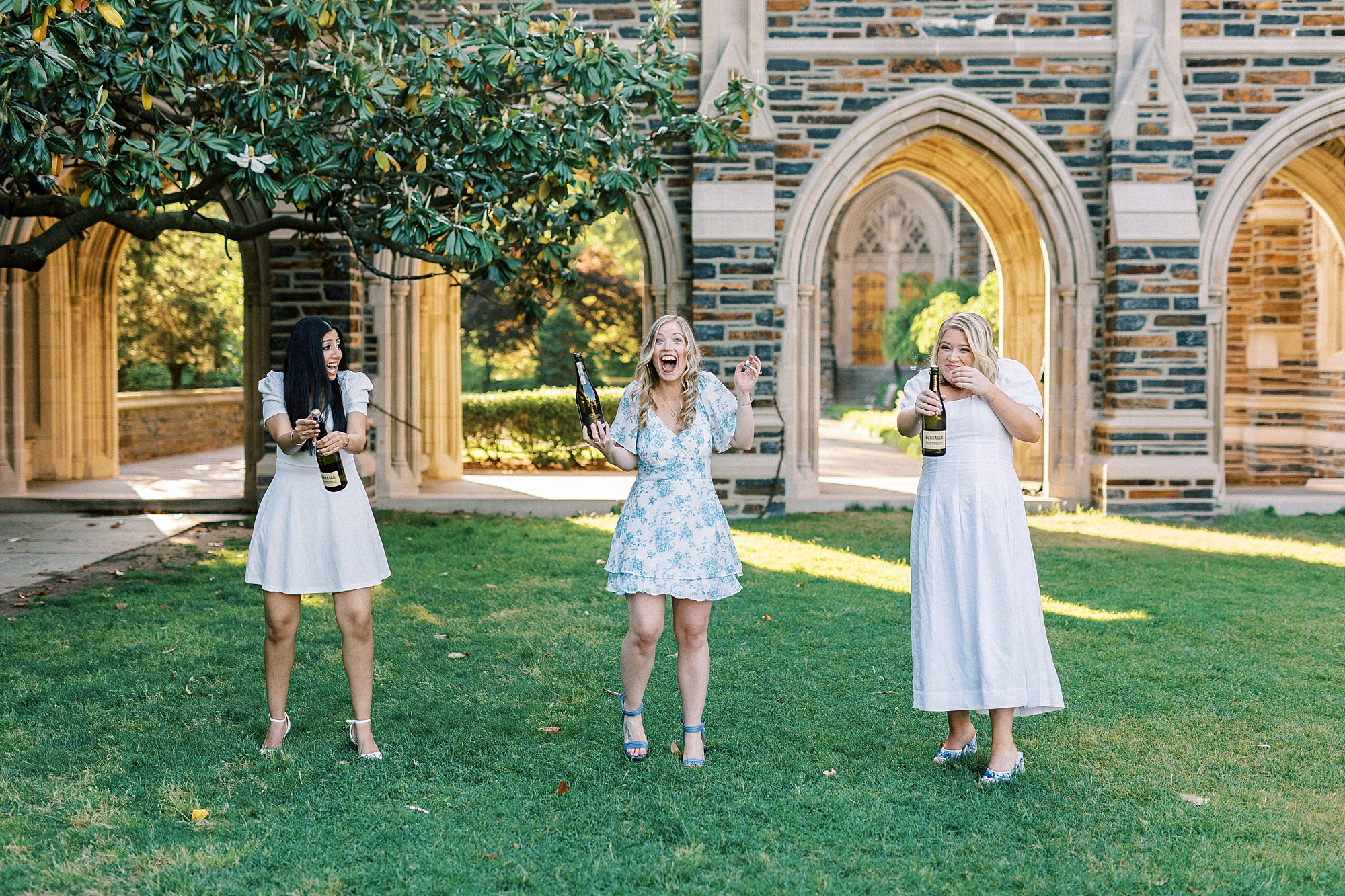 three women pop champagne on lawn outside chapel during graduation portraits at Duke University
